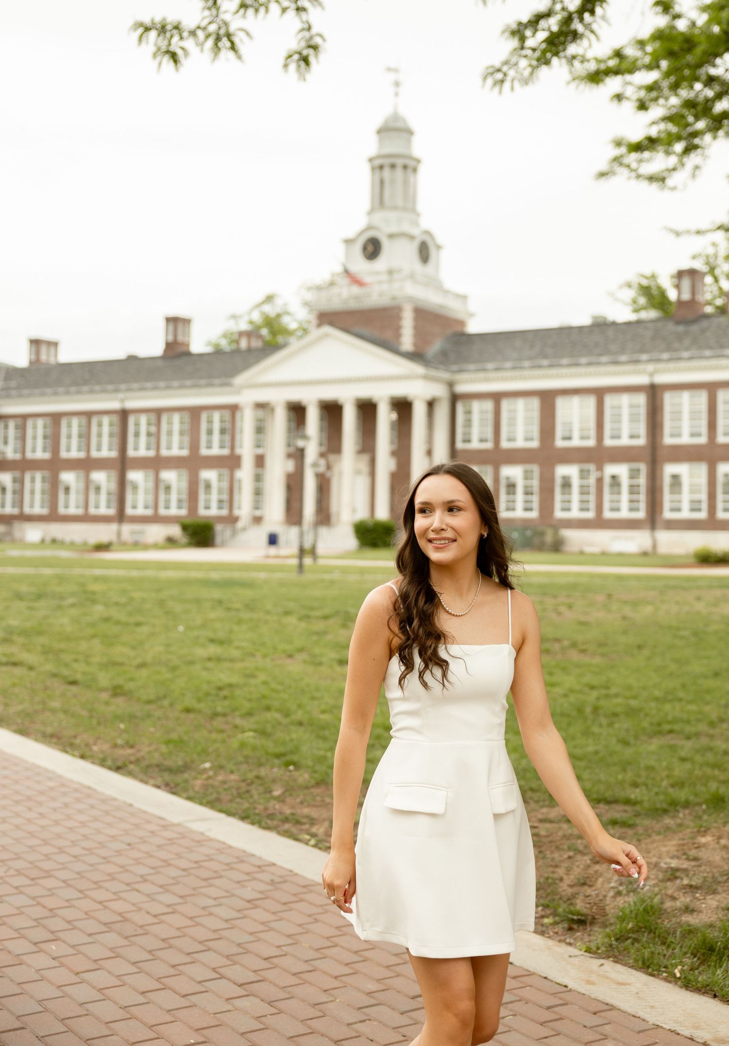 Young woman in white mini dress walking on brick path in front of classic red brick university building.