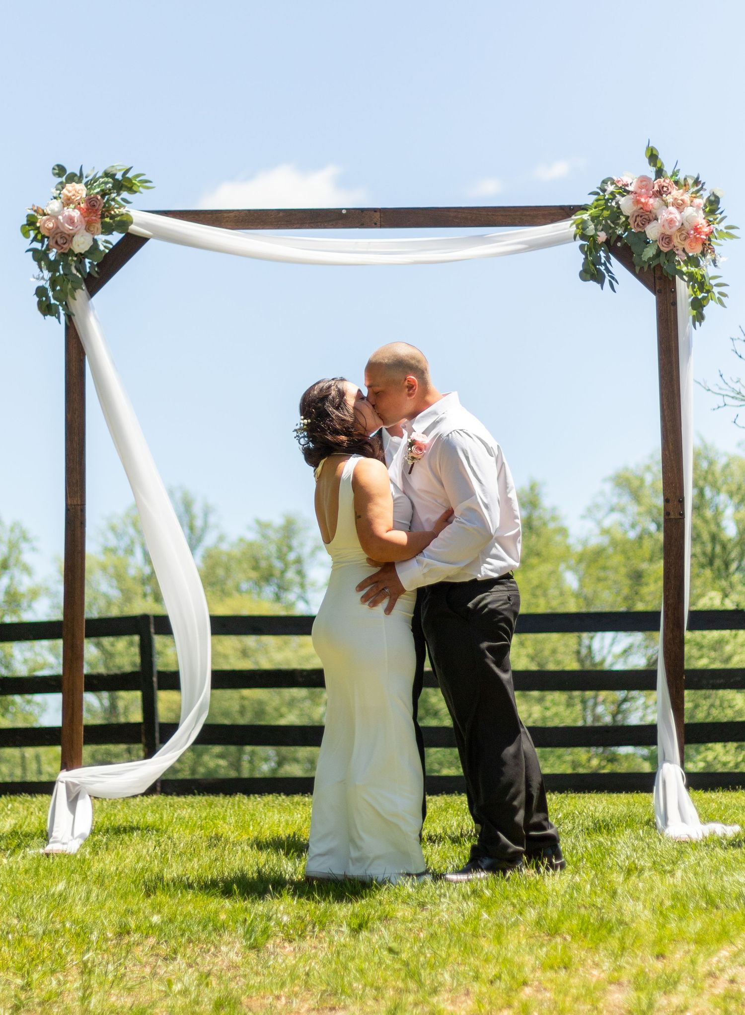 Bride and groom share first kiss under floral wedding arch with white draping on sunny outdoor farm venue.