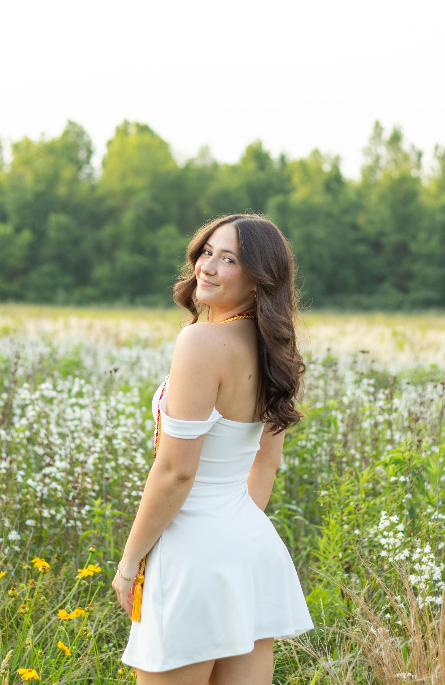 Graduate in white dress poses in field of wildflowers and tall grass.