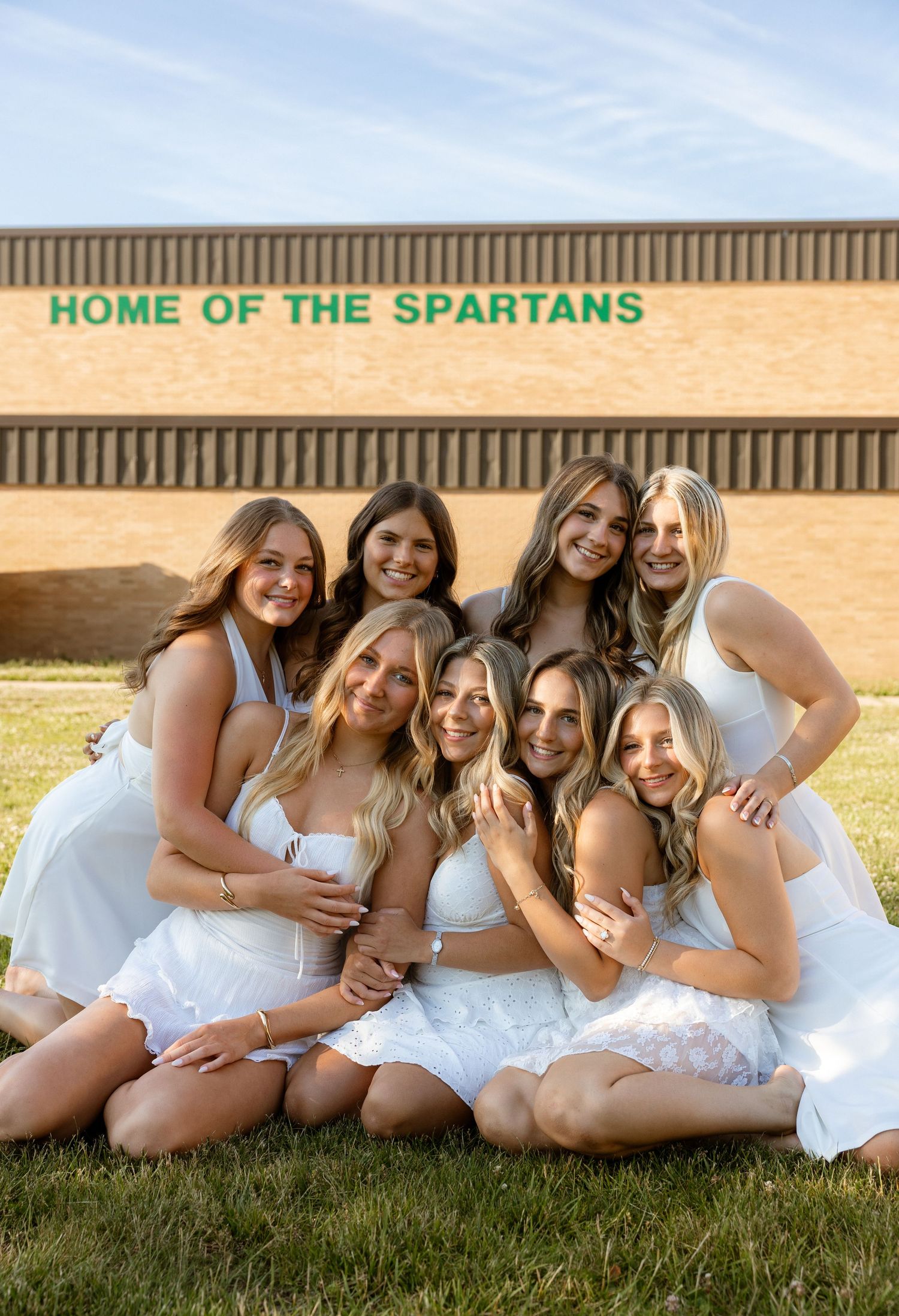 Eight young women in white outfits pose together outdoors in front of a brick building reading Home of the Spartans.