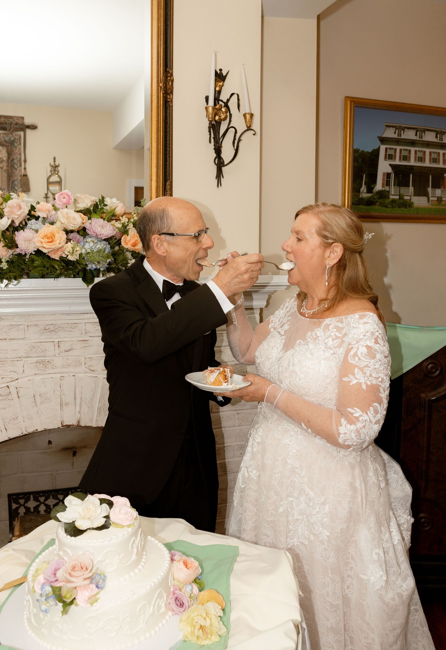 Bride and groom feeding each other wedding cake at reception near fireplace with floral arrangements.