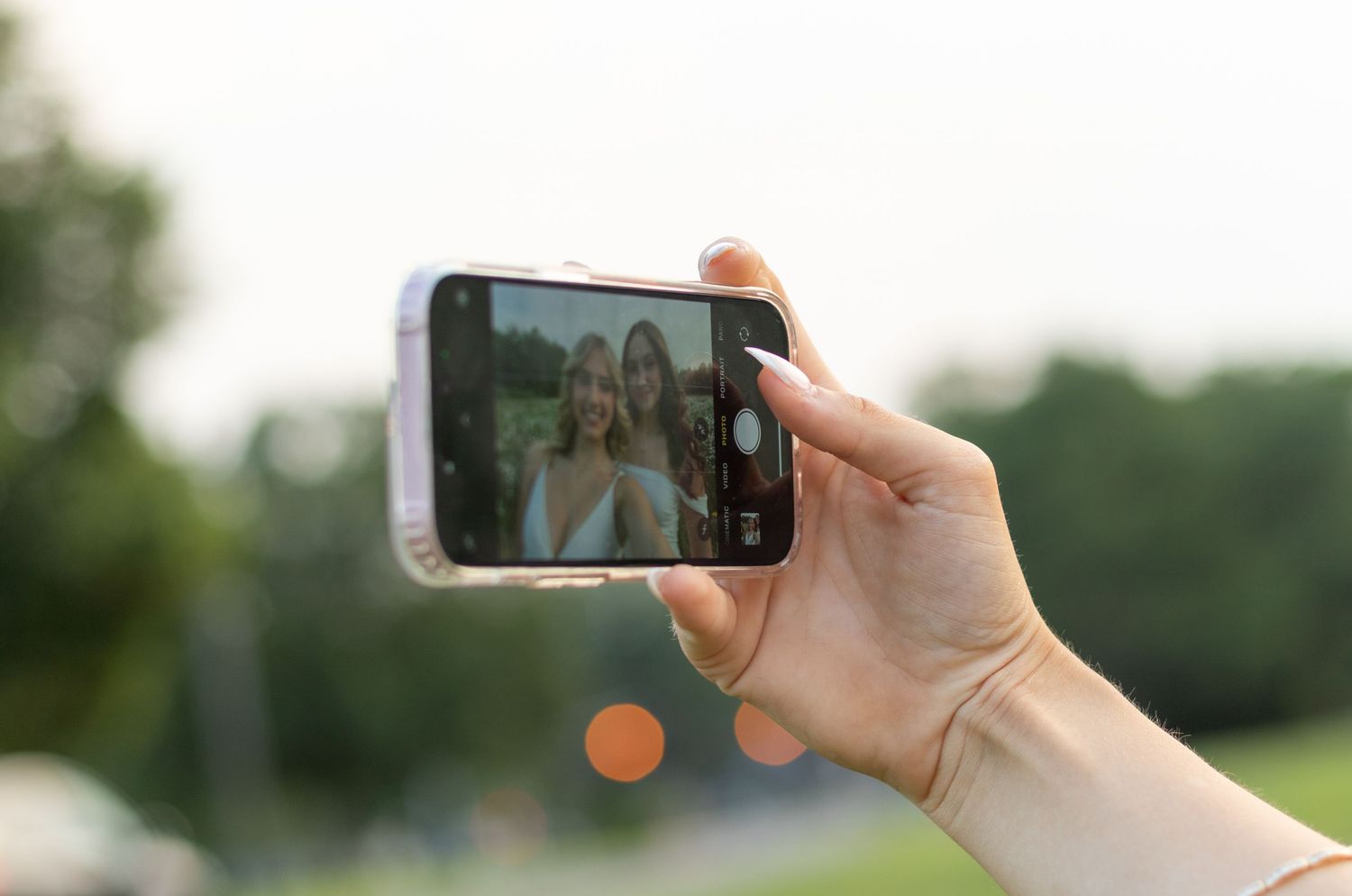 Hand holding smartphone taking graduation photo outdoors at sunset.