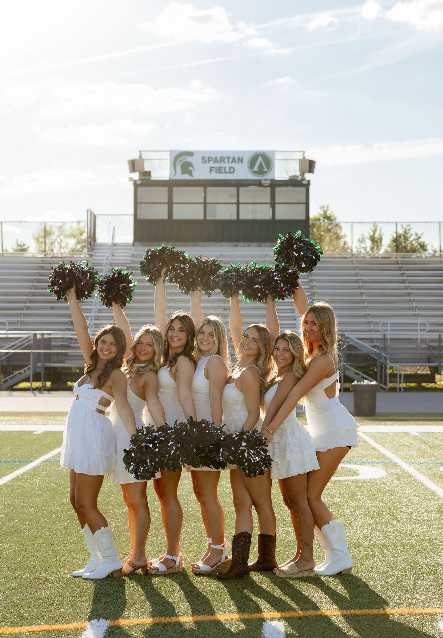 Seven cheerleaders in white uniforms pose with green pom-poms at Spartan Field stadium on a sunny day.