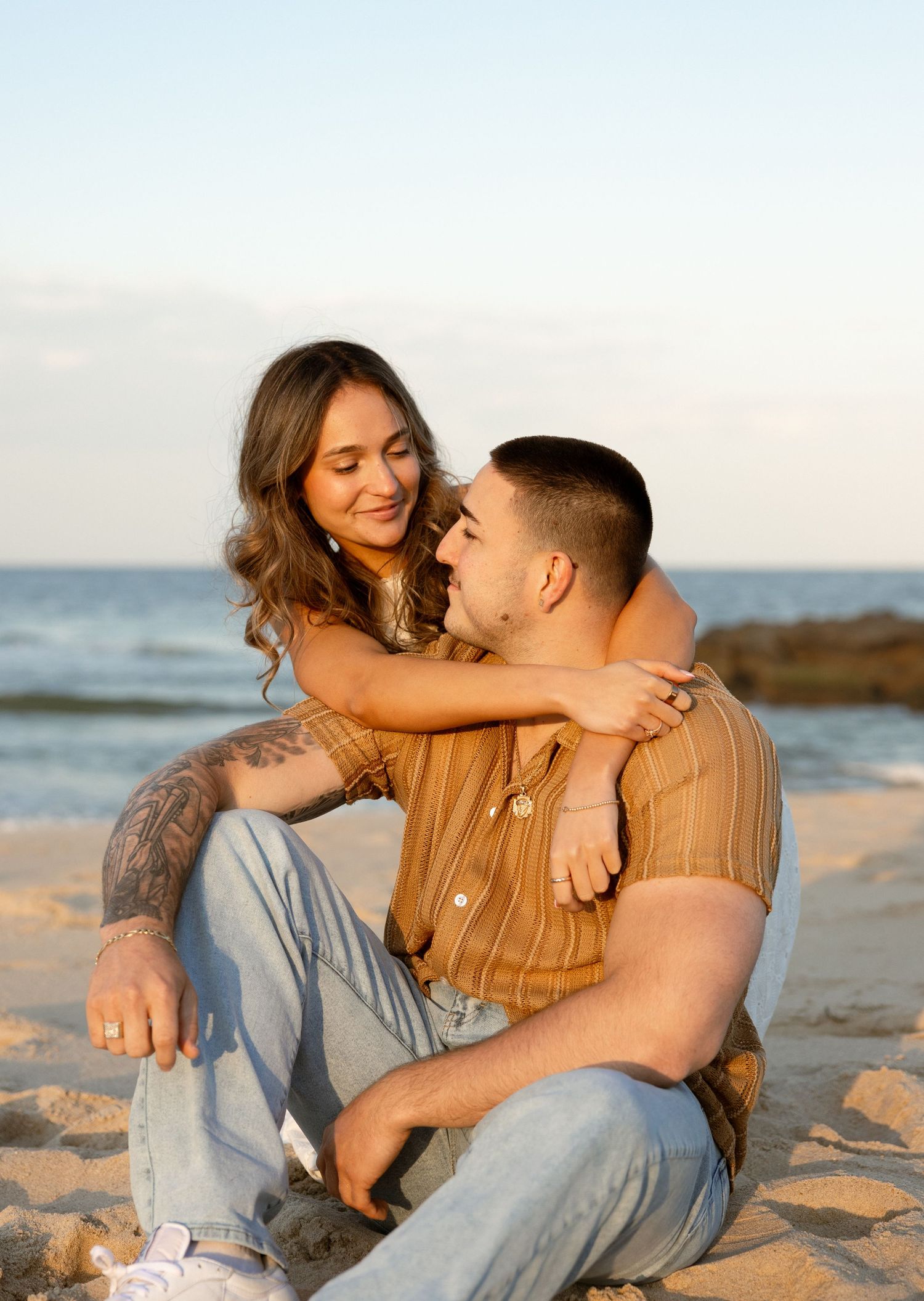 Couple sitting on sandy beach at sunset, woman with tattoos embracing man in striped shirt near ocean.