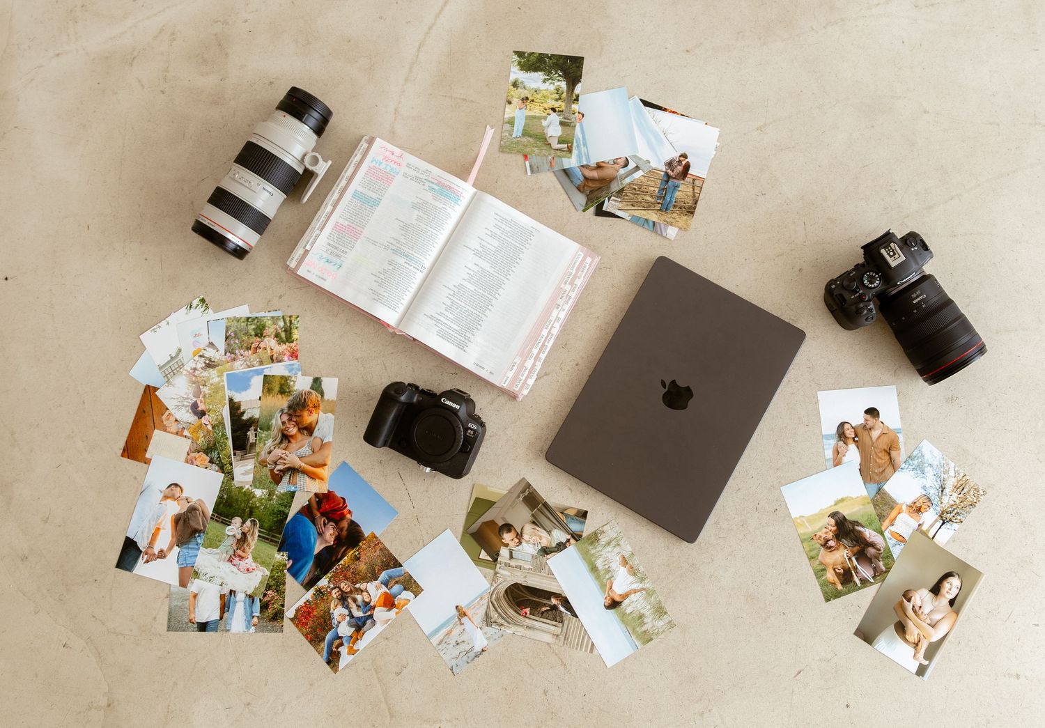 Flat lay of cameras, lenses, an open planner, printed photos, and an Apple MacBook on a beige surface.