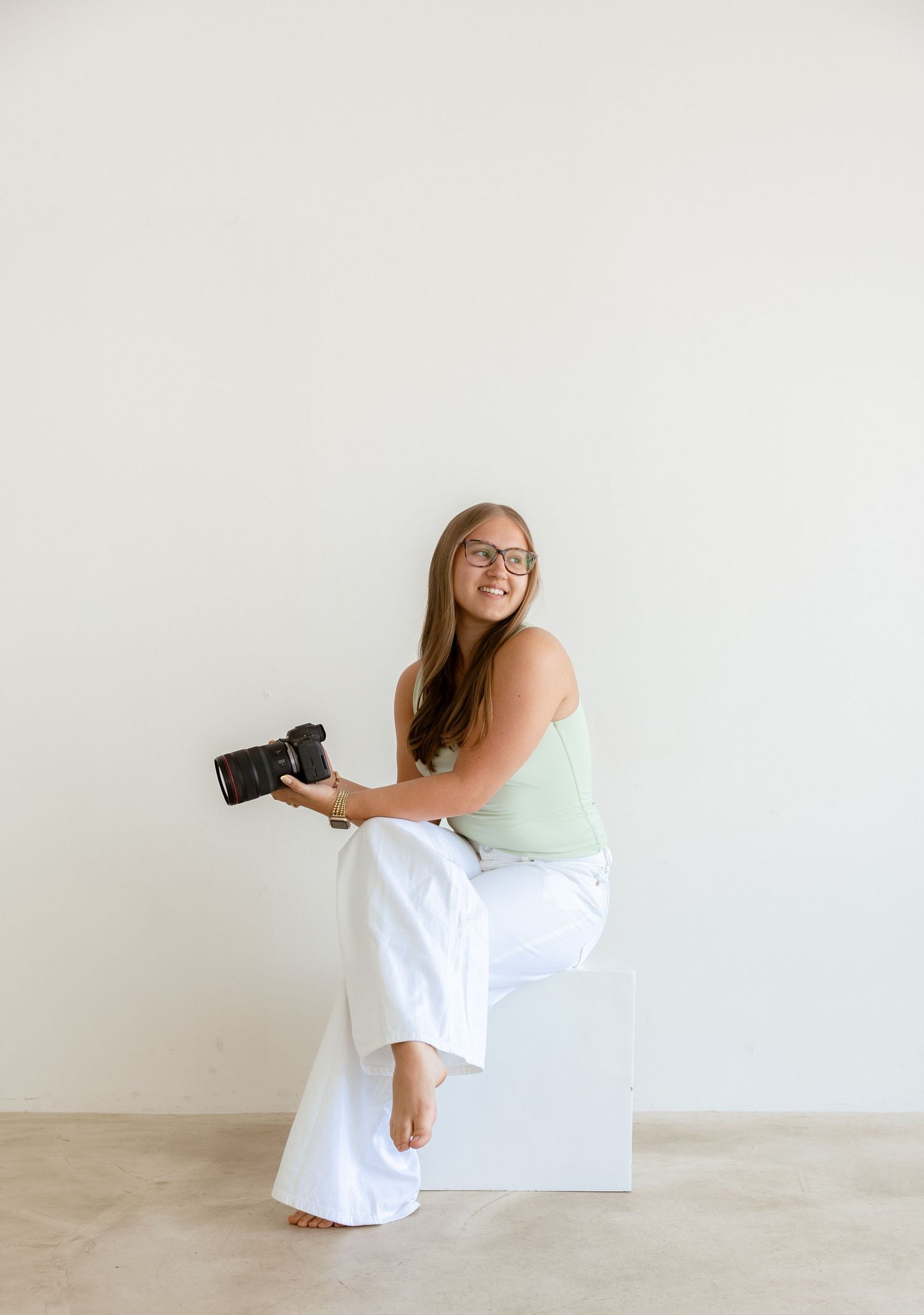 Smiling woman sitting on white cube holding DSLR camera, wearing green tank top and white wide-leg pants.