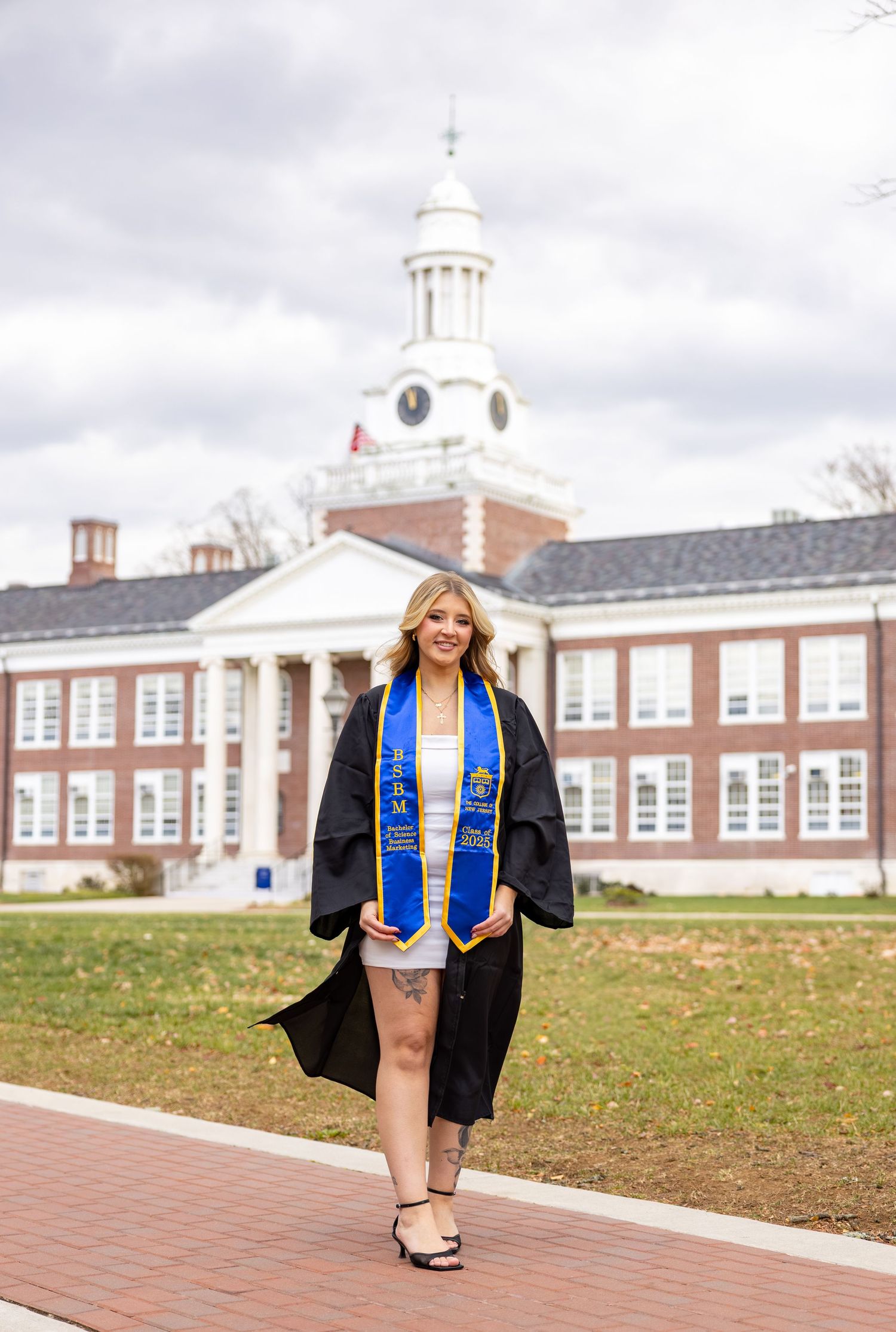 Graduate in cap and gown walking on brick path in front of historic school building with white clock tower.