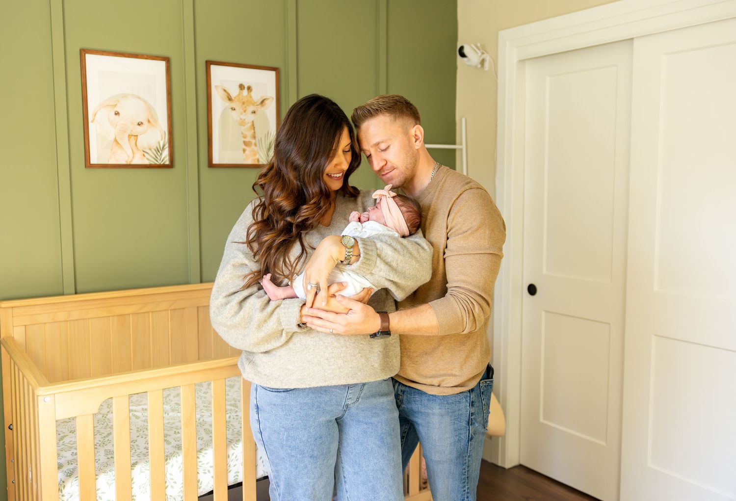 New parents lovingly gaze at their newborn baby in a sage green nursery with a wooden crib.