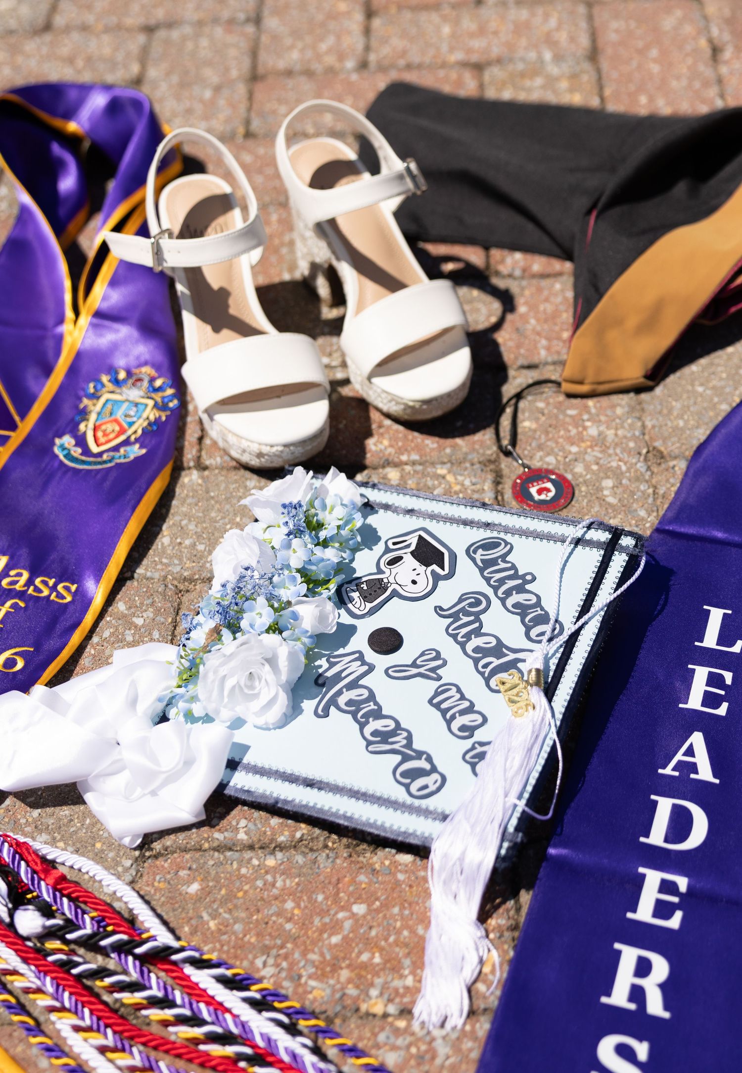 Graduation accessories including white heels, decorated cap, purple sash, honor cords, and leadership stole on brick pavement.