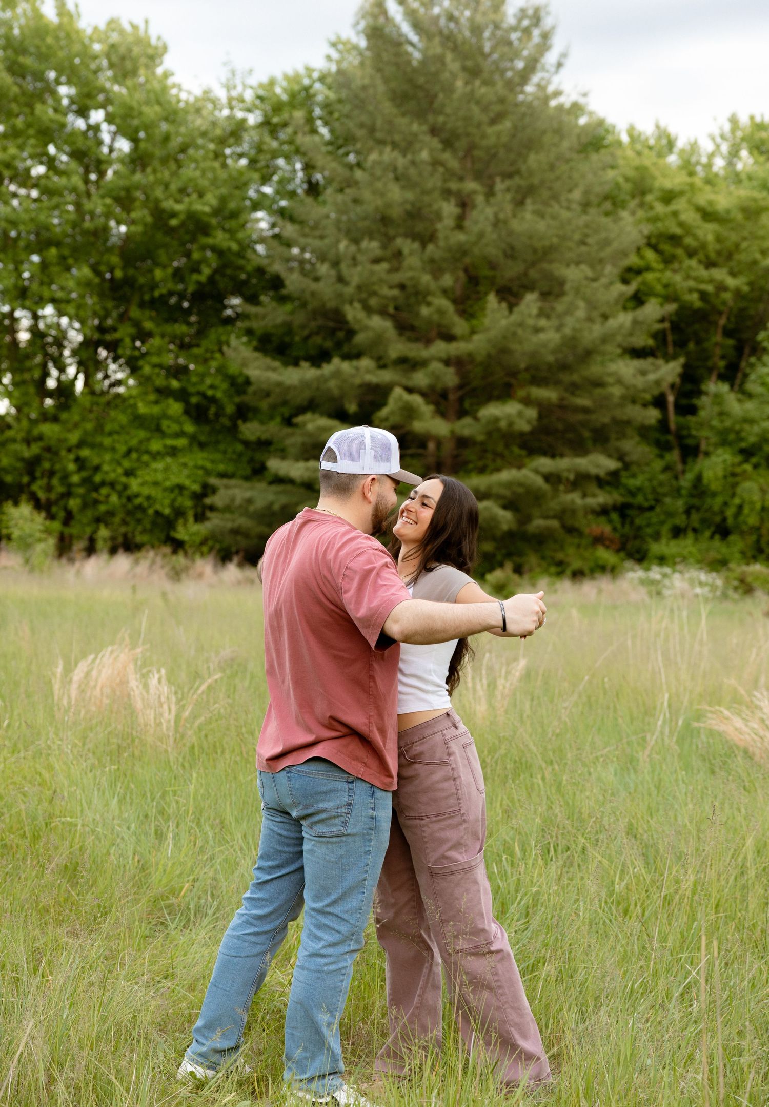 A couple dances and embraces in a lush green meadow surrounded by tall trees on a cloudy day.