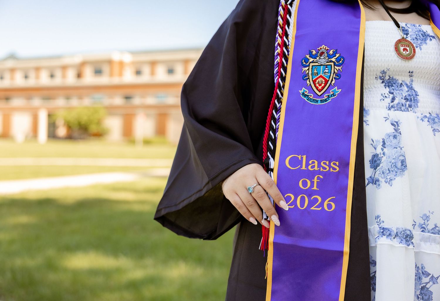 Graduate wearing black robe with purple Class of 2026 honor sash on college campus lawn.