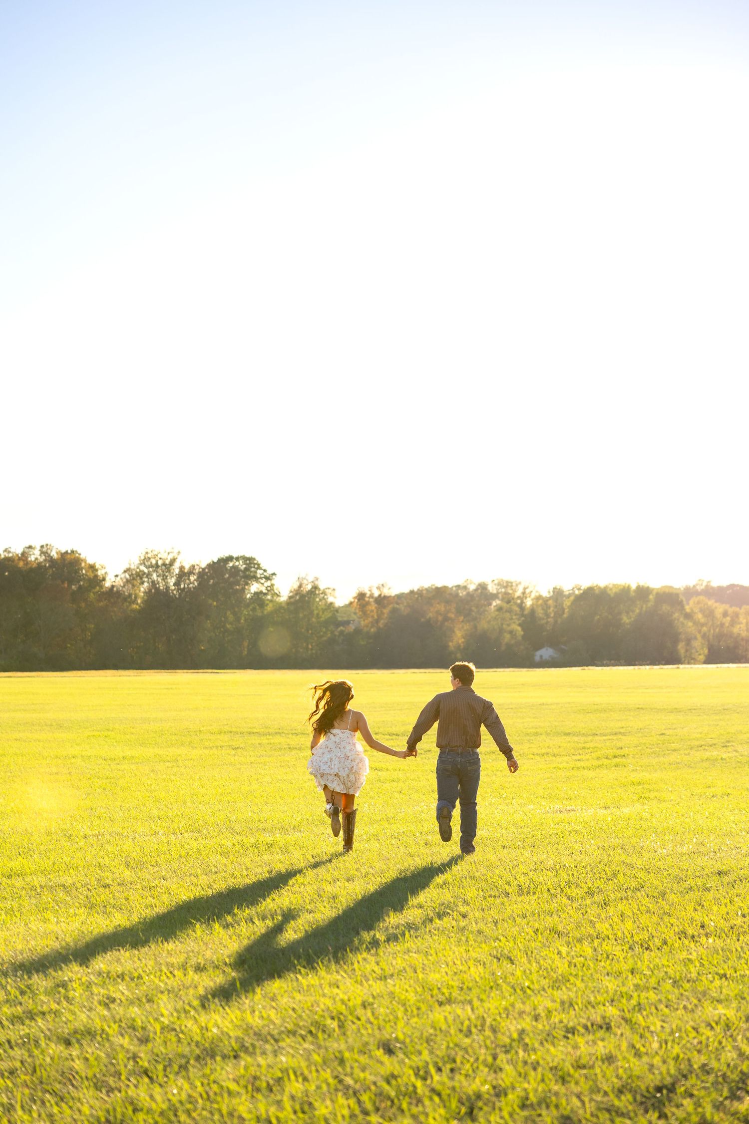 A couple walks hand in hand across a sunlit meadow casting long shadows at sunset.