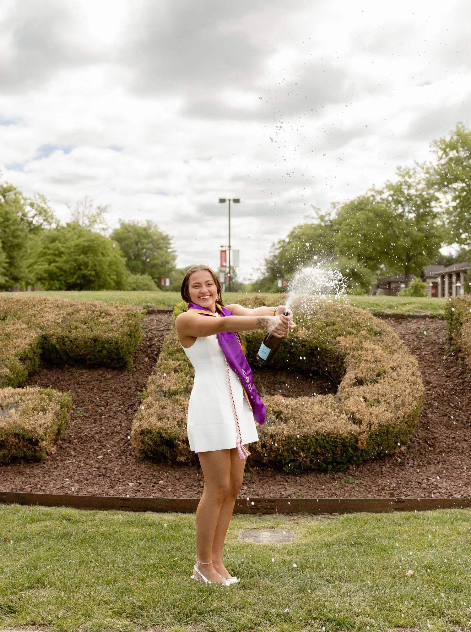 Young woman in white dress and purple sash celebrates graduation by popping champagne bottle outdoors near decorative hedges.