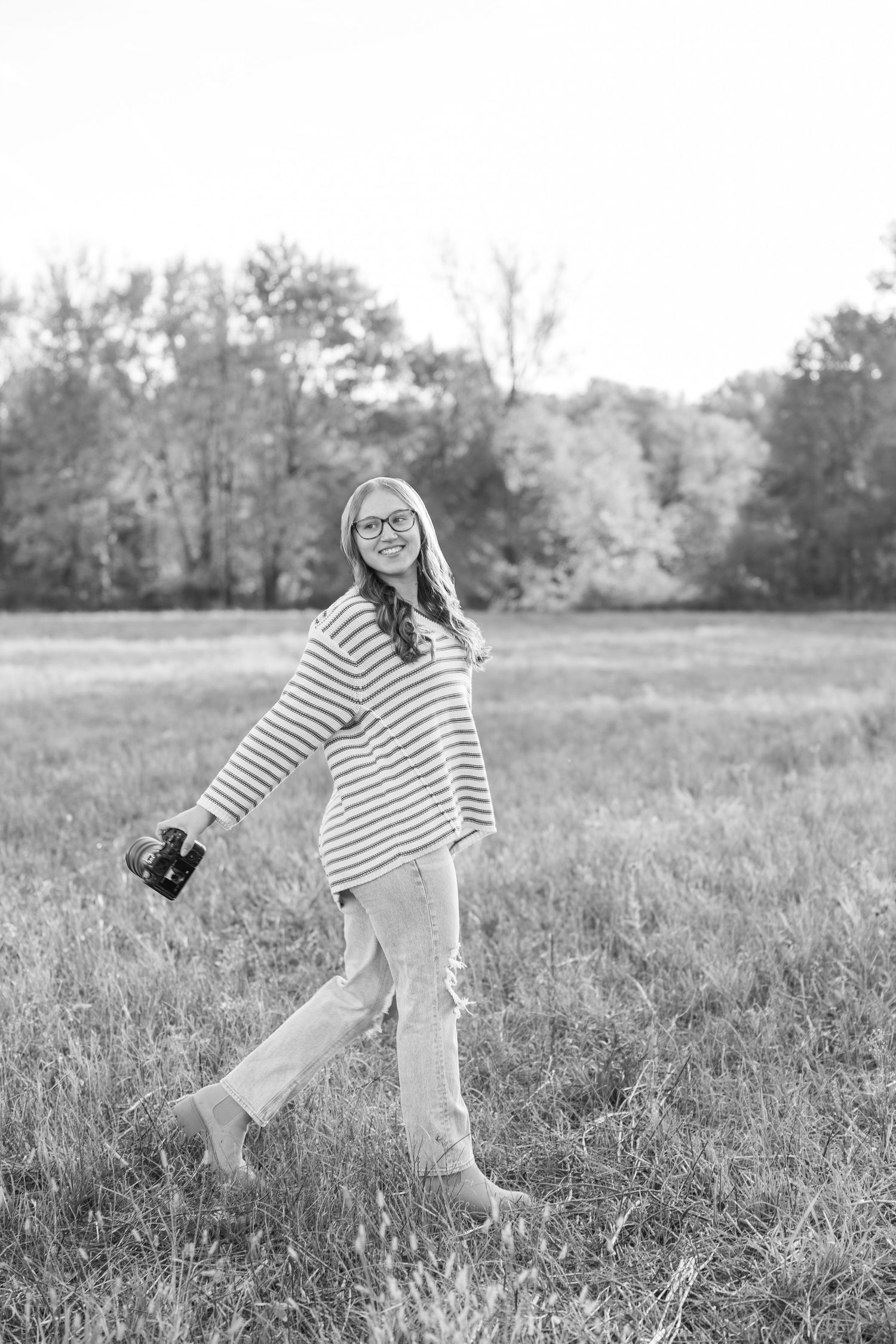 Person in striped shirt walking through field holding camera in black and white.