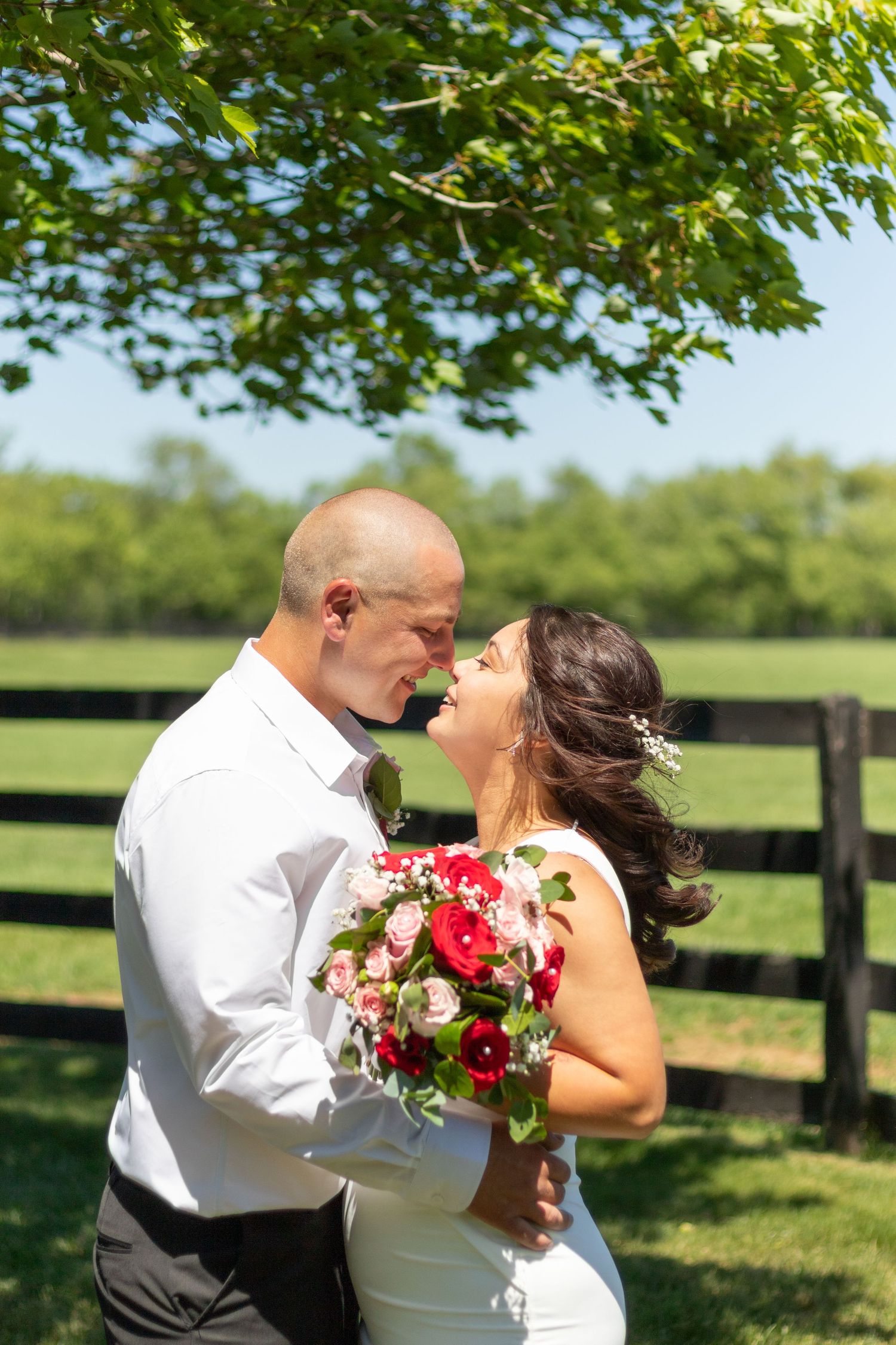 Newlyweds embrace in sunny field with red and white bouquet near wooden fence.