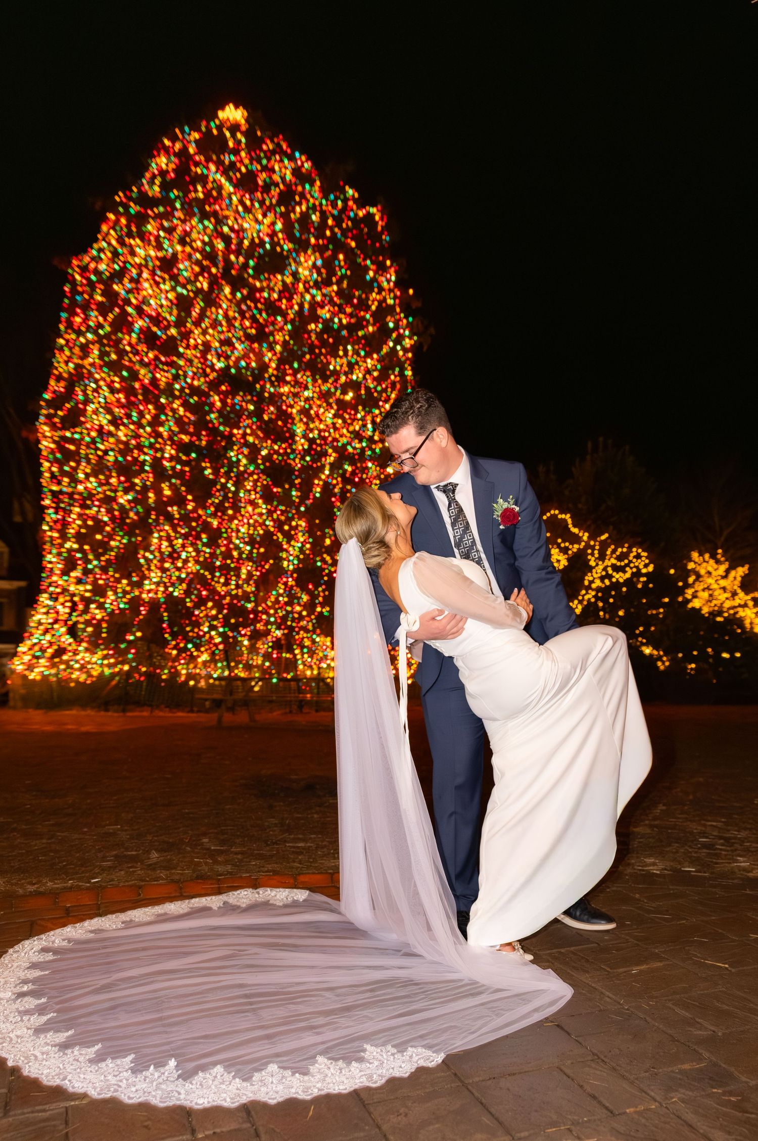 Newlyweds dance under illuminated trees at night with flowing white wedding dress.