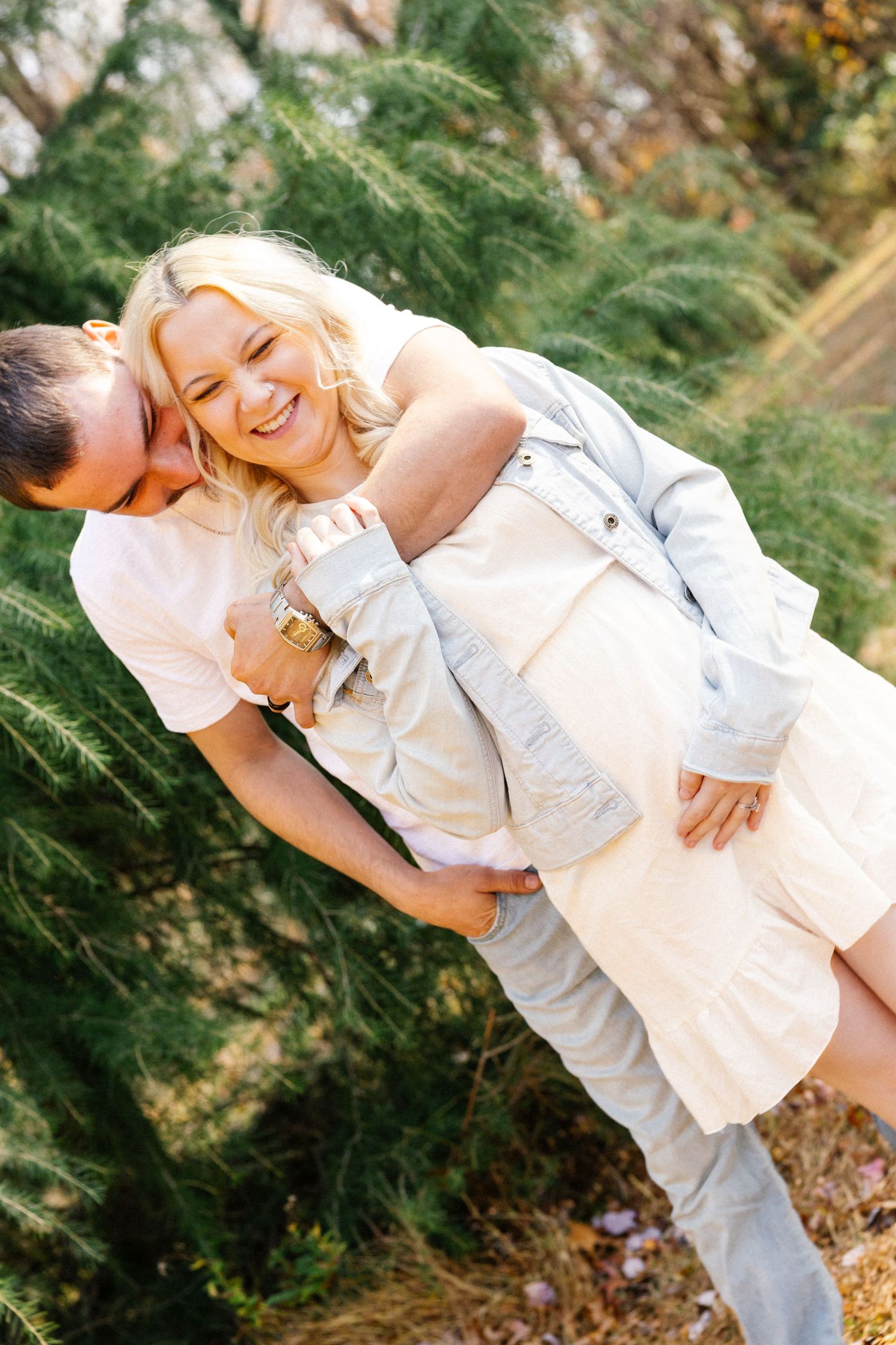 A joyful outdoor portrait of a couple in light-colored clothing against evergreen trees.