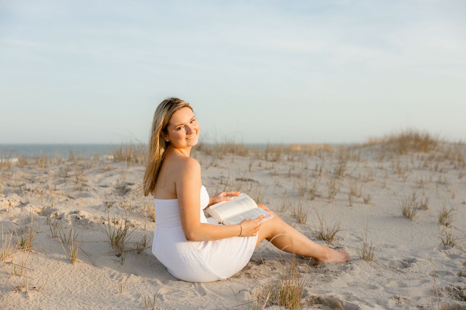 Young woman in white dress sitting on sandy beach dunes, smiling over shoulder at golden hour sunset.