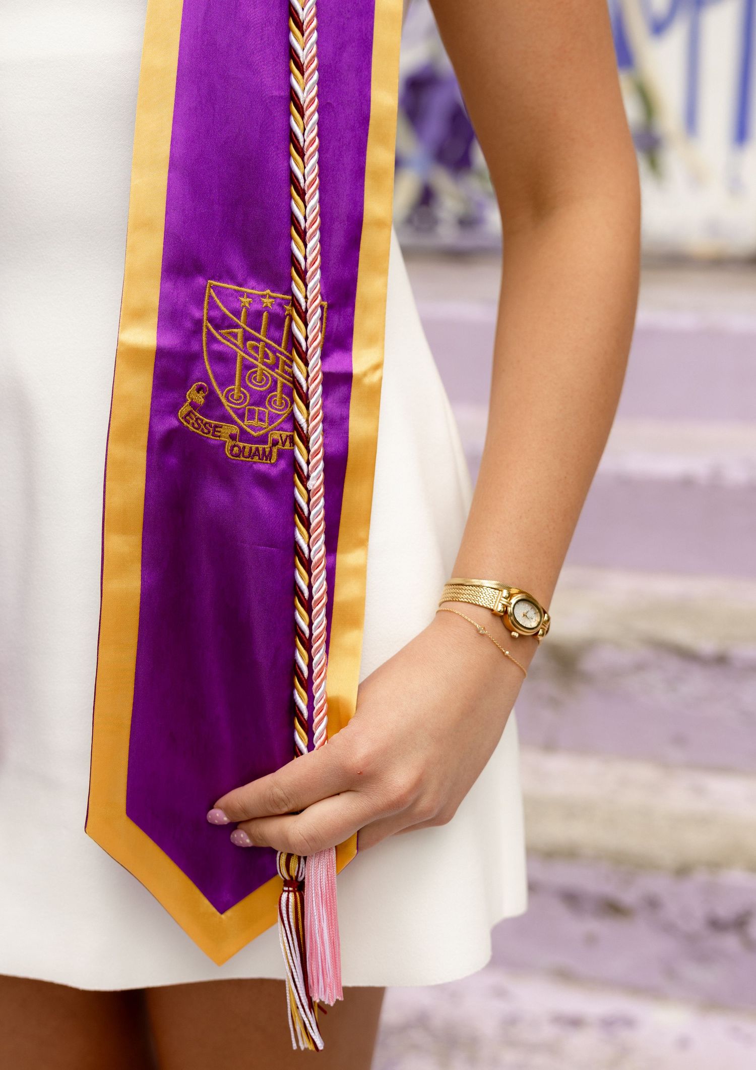 Woman in white dress holding purple and gold graduation stole with crest, wearing gold bracelet and watch.