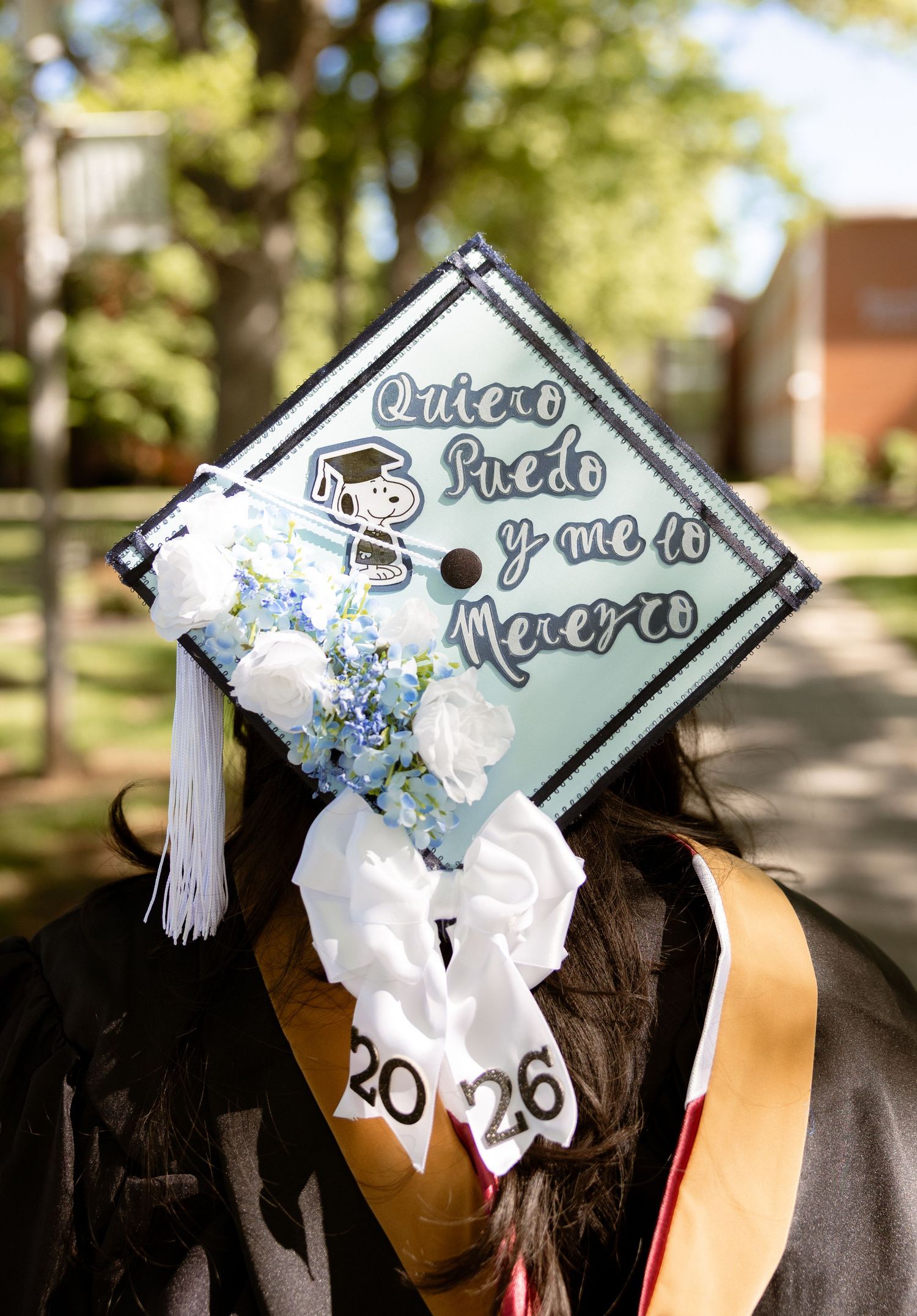 Graduate wearing decorated mortarboard cap with Snoopy and Spanish text 'Quiero Puedo y me lo Merezco' at commencement.