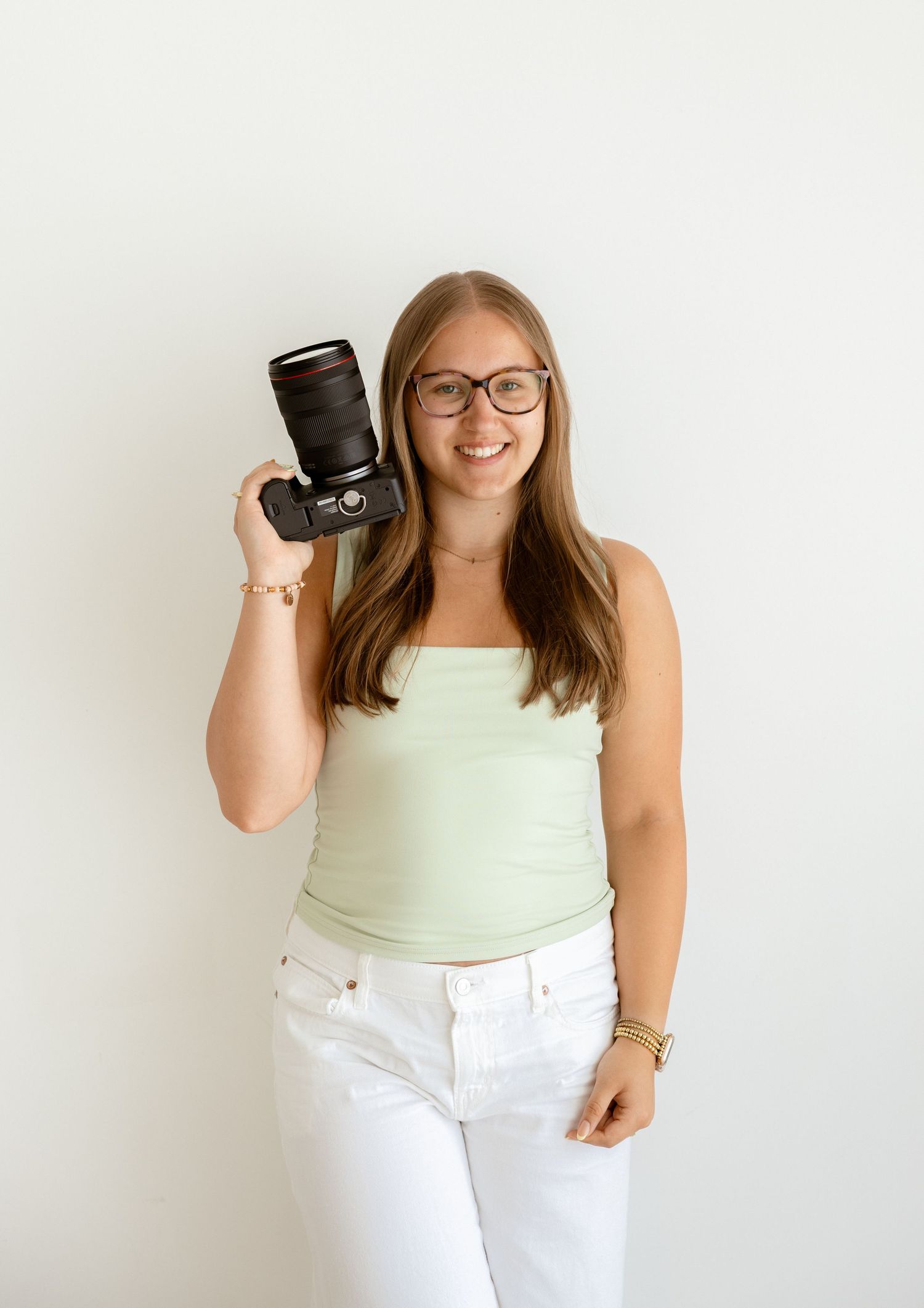 Smiling young woman with glasses holding a DSLR camera, wearing a mint top and white jeans against a white background.
