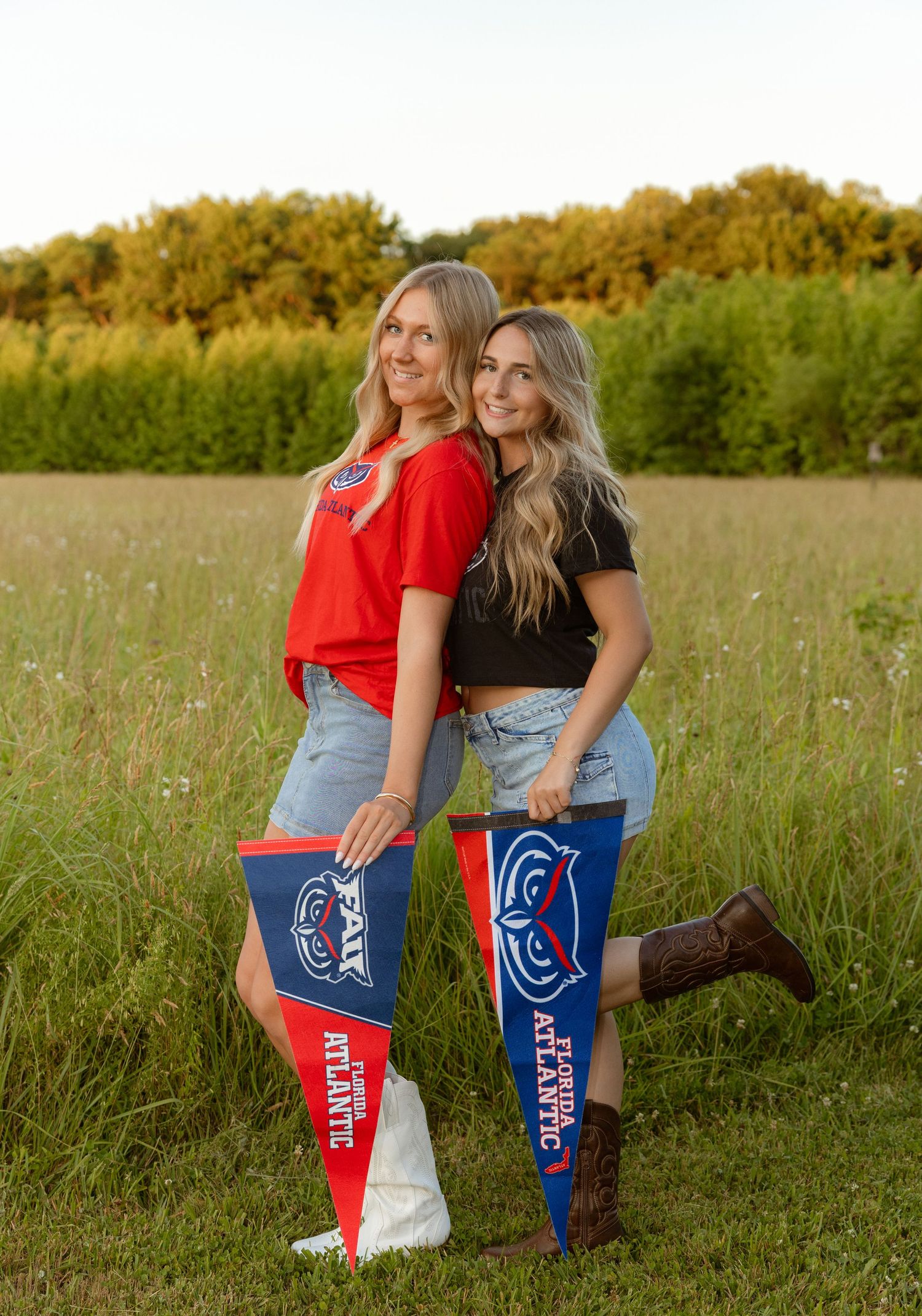 Two smiling women holding Florida Atlantic University pennants pose in a sunny field with green trees in the background.