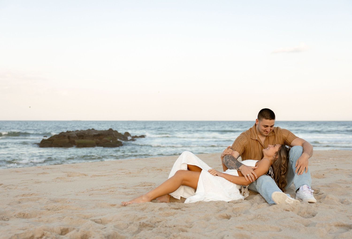 Romantic couple relaxing together on a sandy beach at sunset, with ocean waves and rocks in the background.