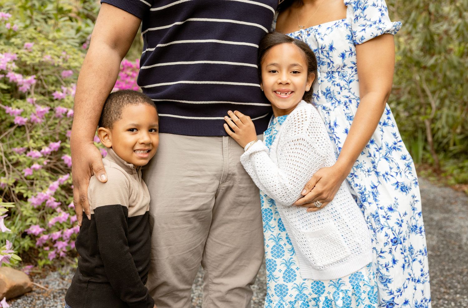 Family of four posing outdoors near pink flowers, two children smiling beside their parents on a garden path.
