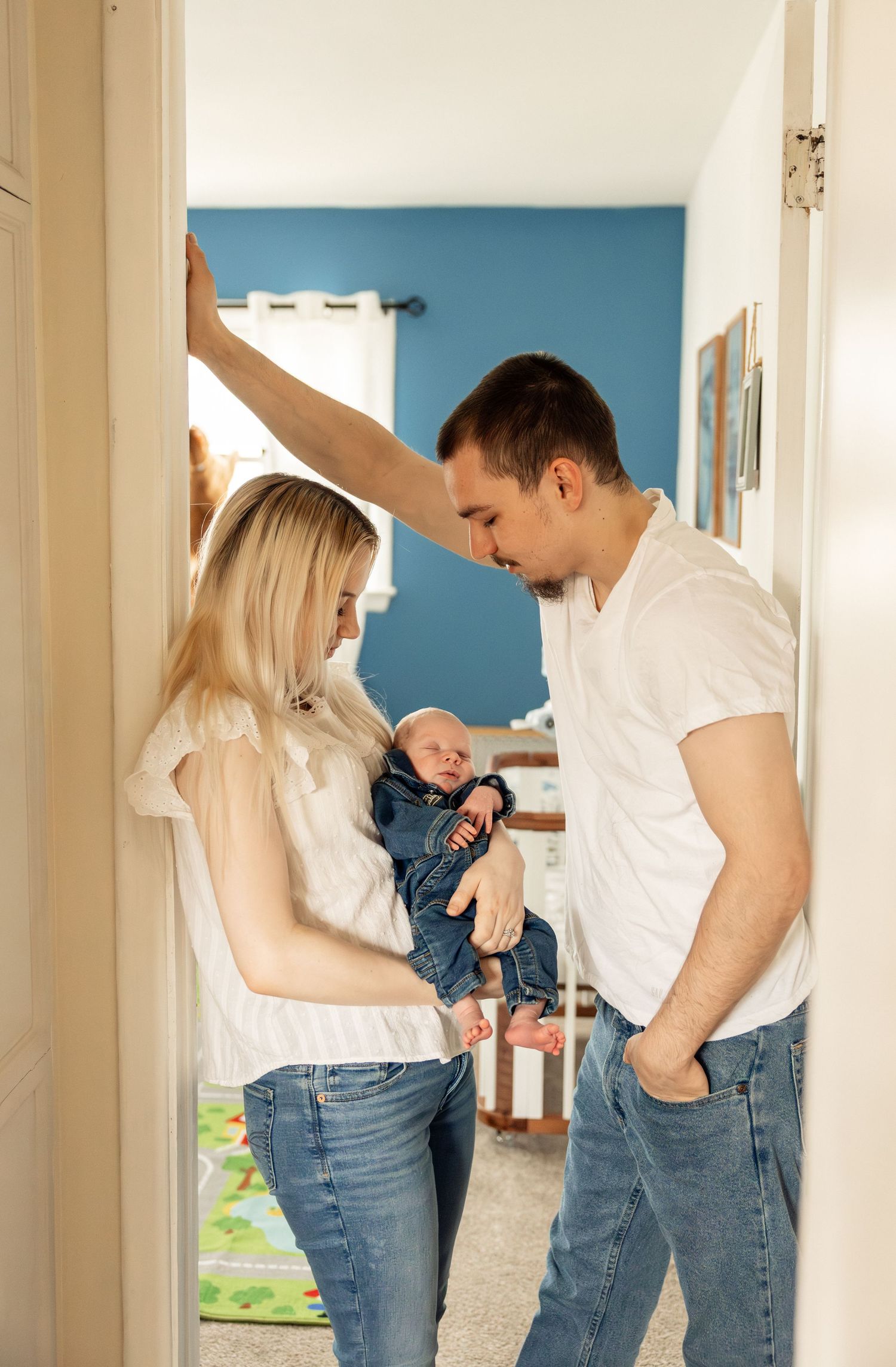 Young couple in white tops and jeans lovingly gazing at their newborn baby in a cozy home nursery.