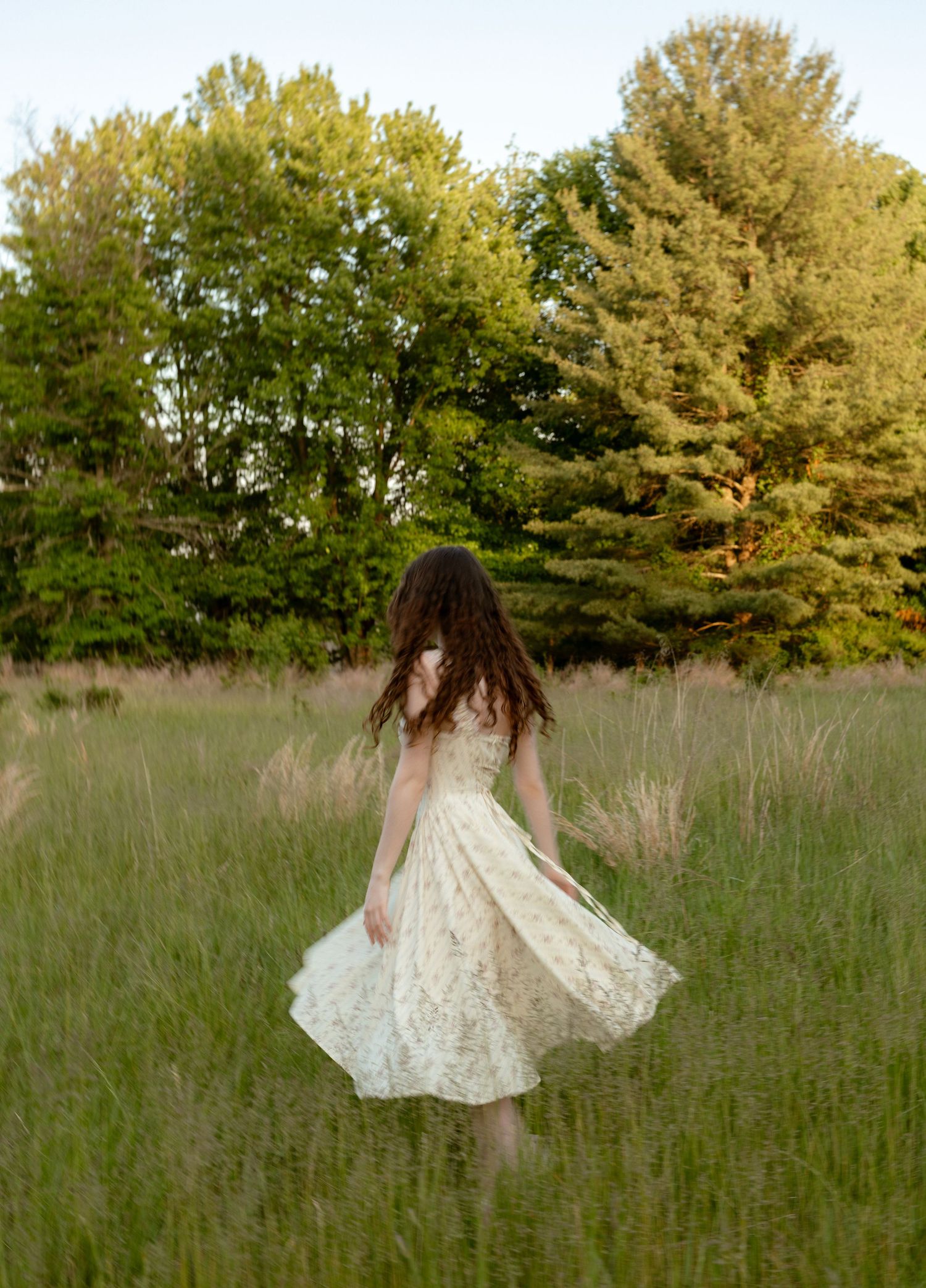 A young woman in a white lace dress twirls in a lush green meadow surrounded by tall trees at golden hour.