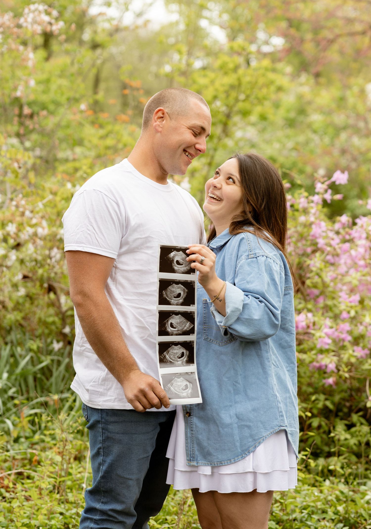 Happy couple holding ultrasound photos outdoors among blooming pink flowers, celebrating pregnancy news together.