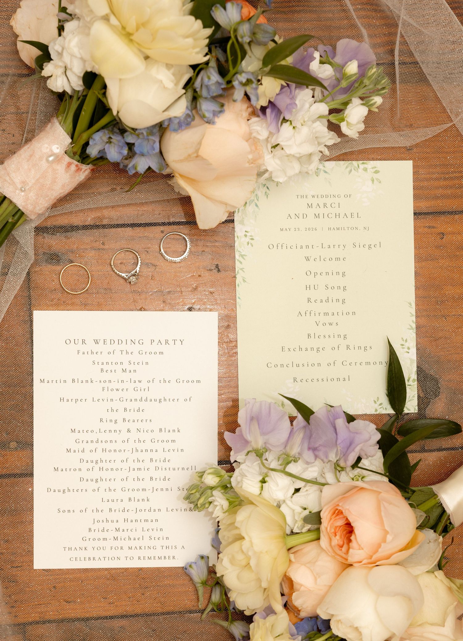 Elegant wedding stationery suite with floral bouquets featuring peach roses, blue flowers, and gold rings on a rustic wood surface.