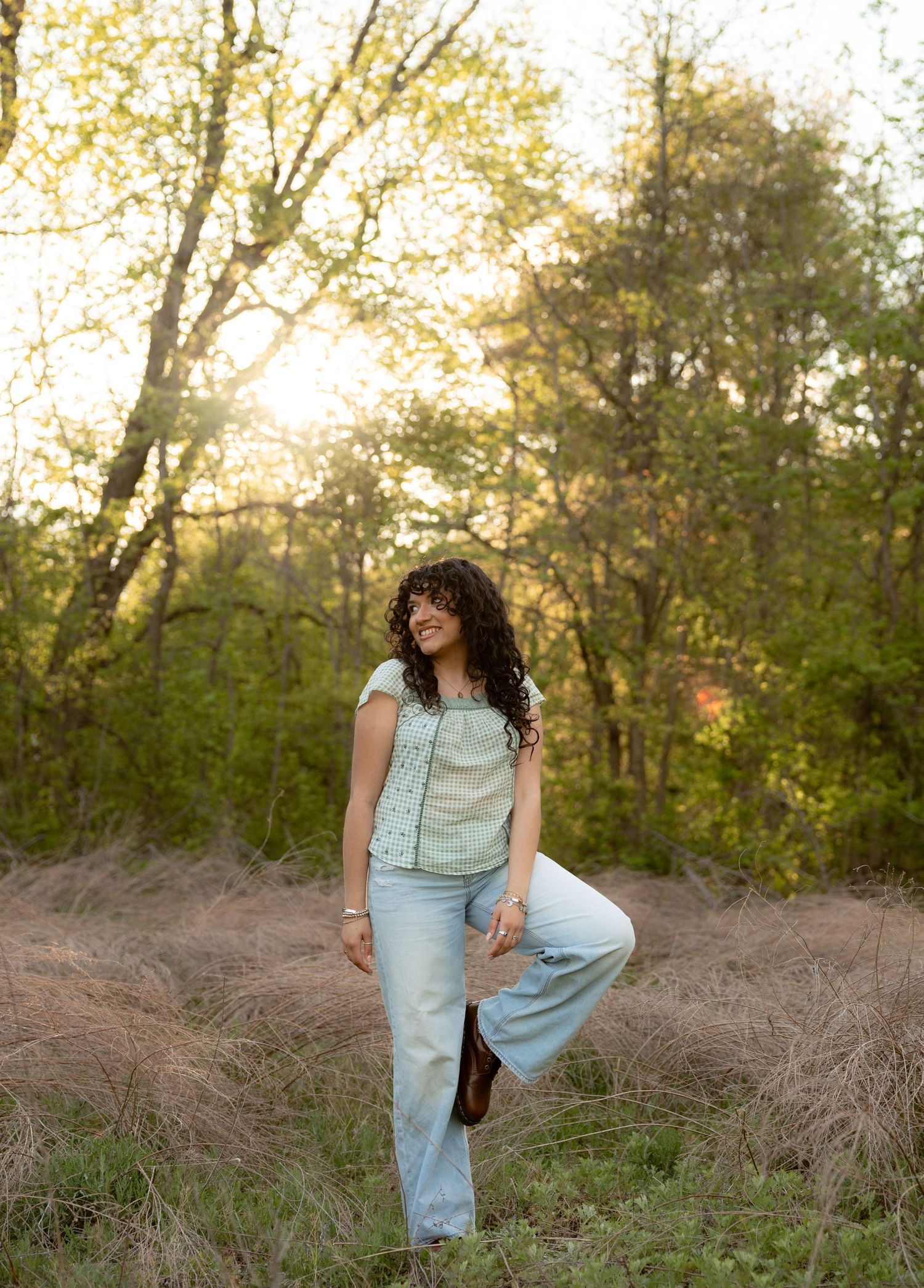Woman in light blue jeans and top standing in sunlit meadow with trees, golden hour light.