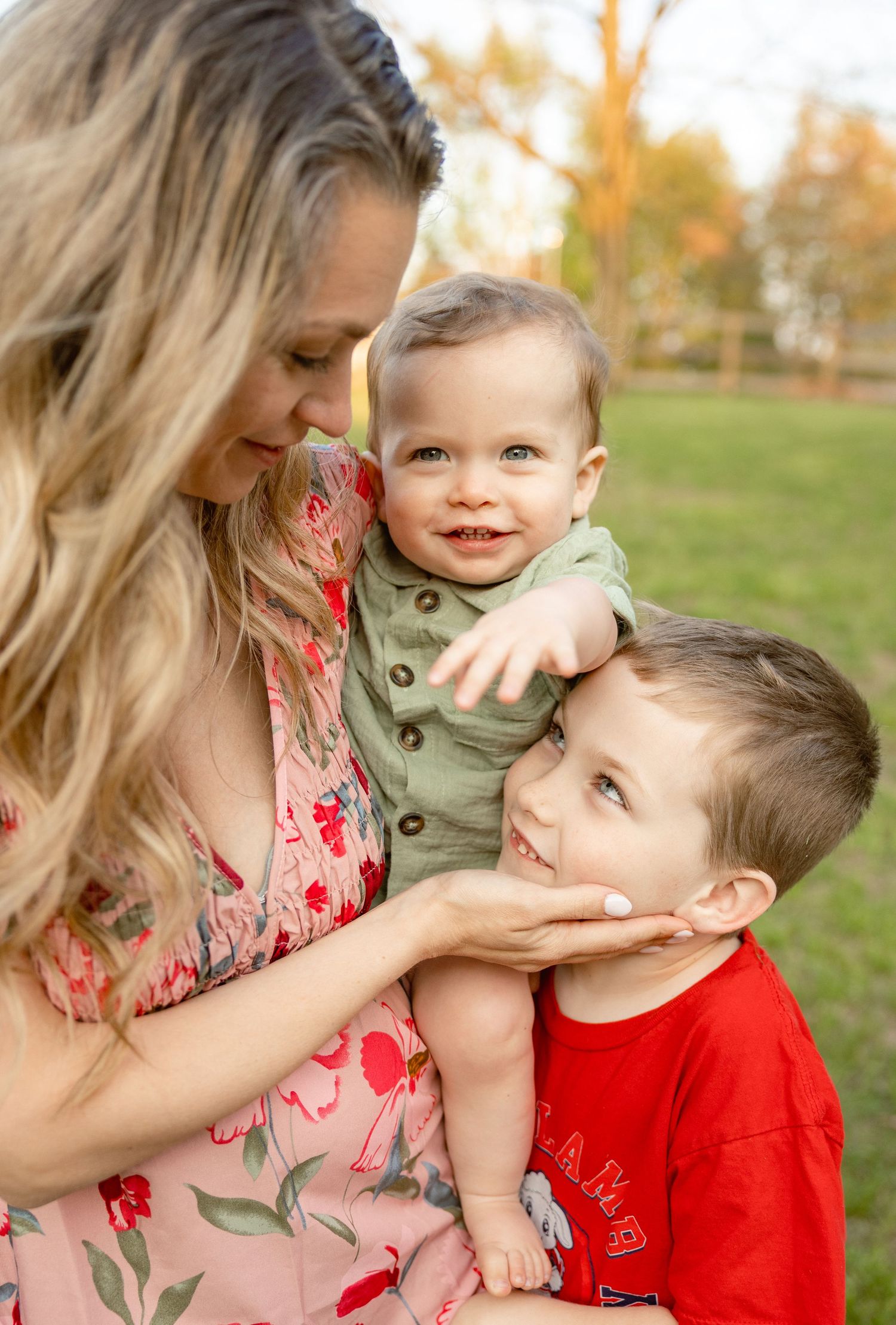 A smiling mother holds a happy baby and young boy in a red shirt outdoors in a park during fall.
