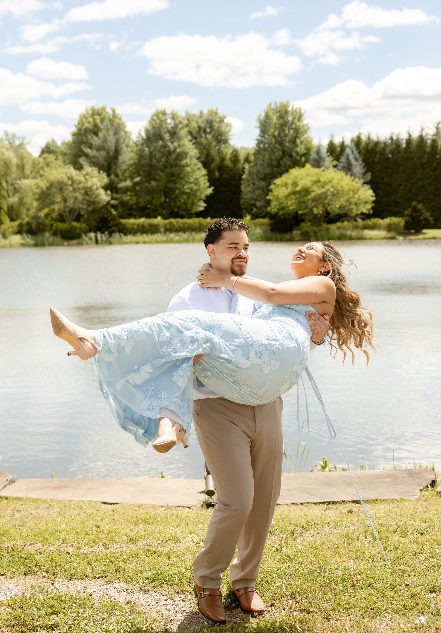 A man joyfully carries a woman in a light blue dress by a serene lake on a sunny day.
