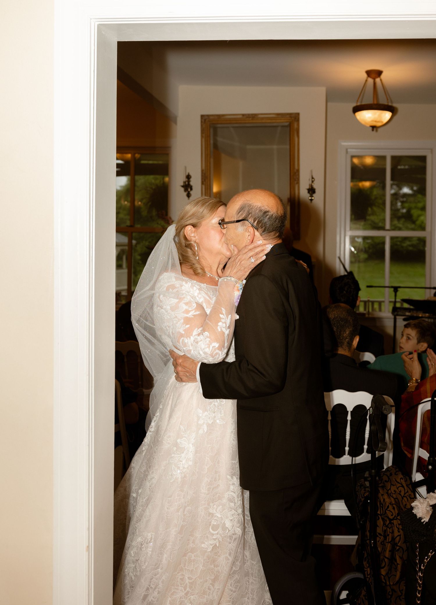 Elderly bride and groom share a kiss at their wedding reception, bride wearing lace gown and veil.
