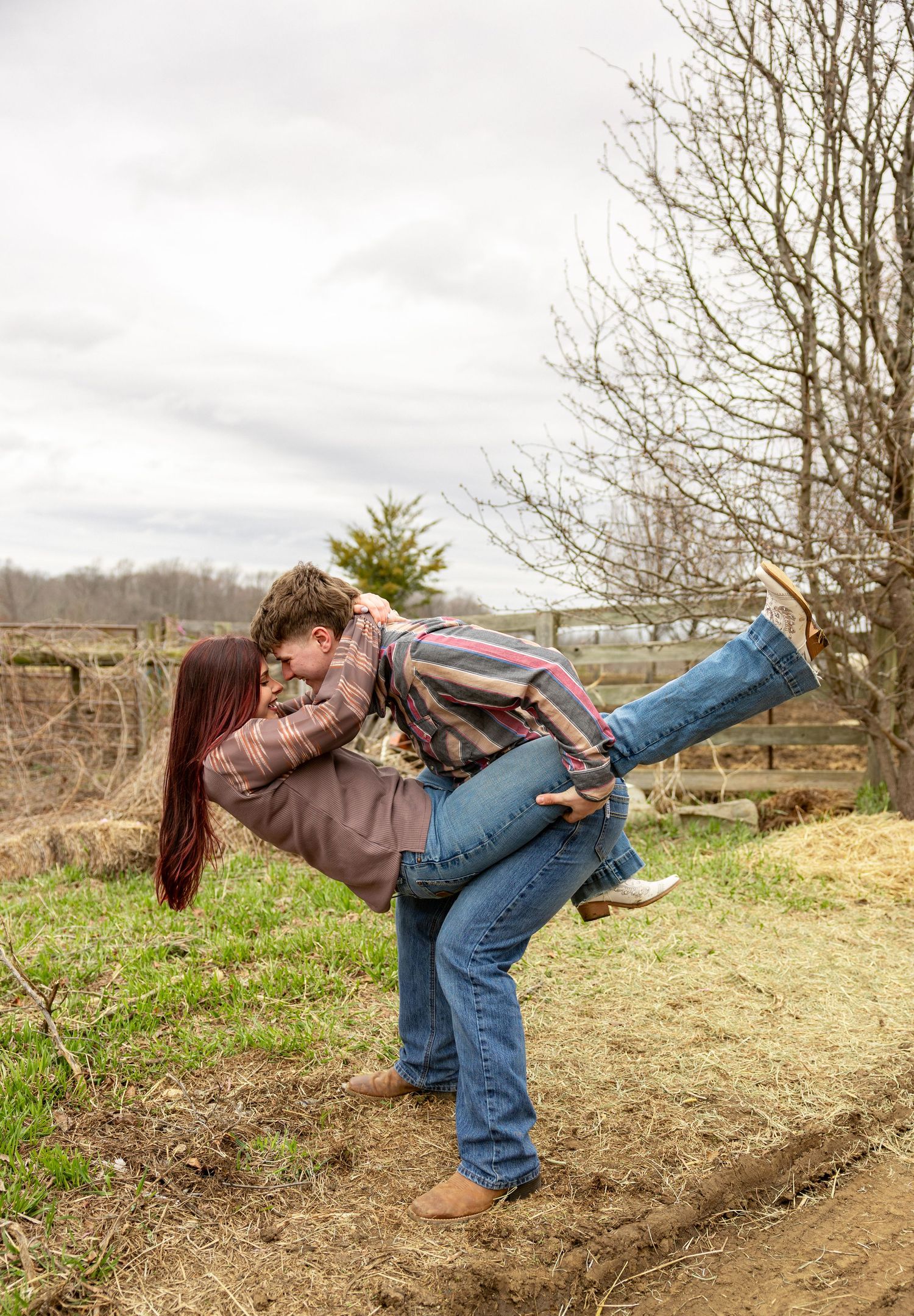 Couple sharing a romantic dip kiss outdoors on a farm, surrounded by bare trees and green grass.