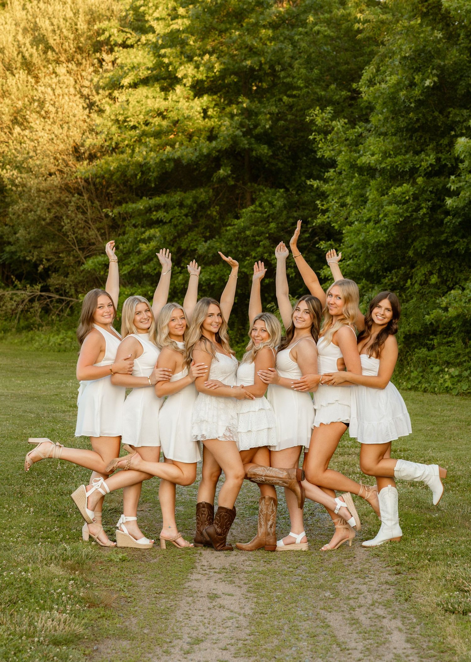 Group of nine women in white dresses and boots celebrating outdoors on a sunny day in a green field.