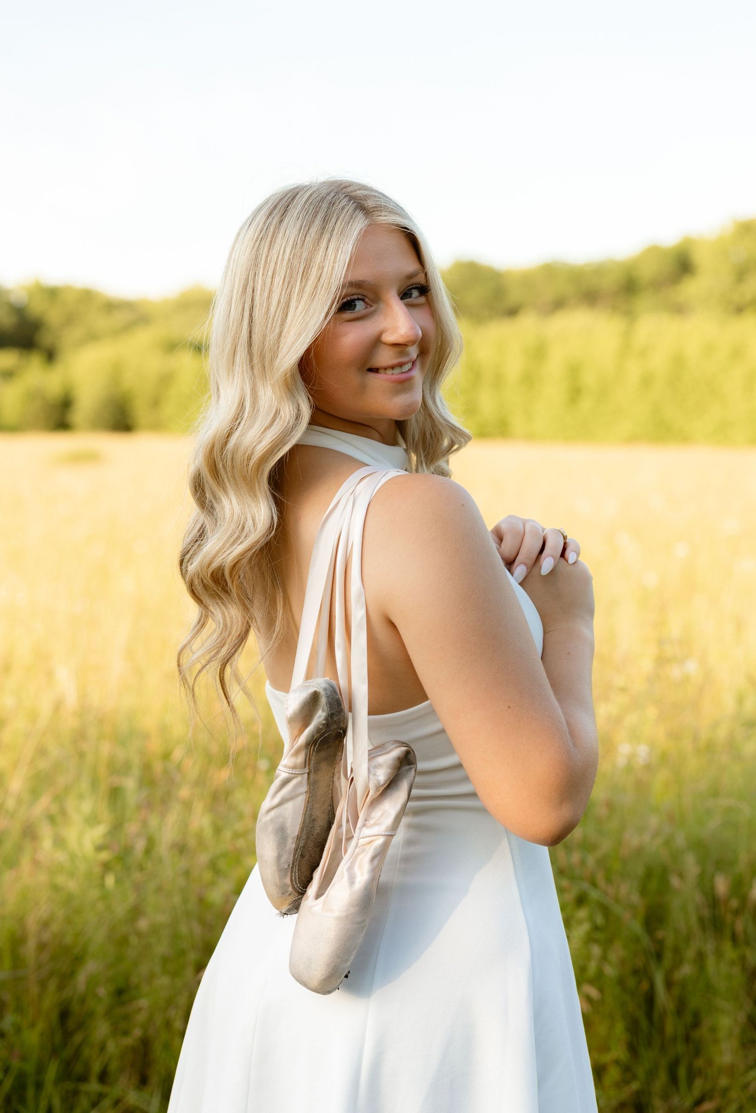 Blonde teenage girl in white dress holding ballet pointe shoes over shoulder in golden sunlit field.