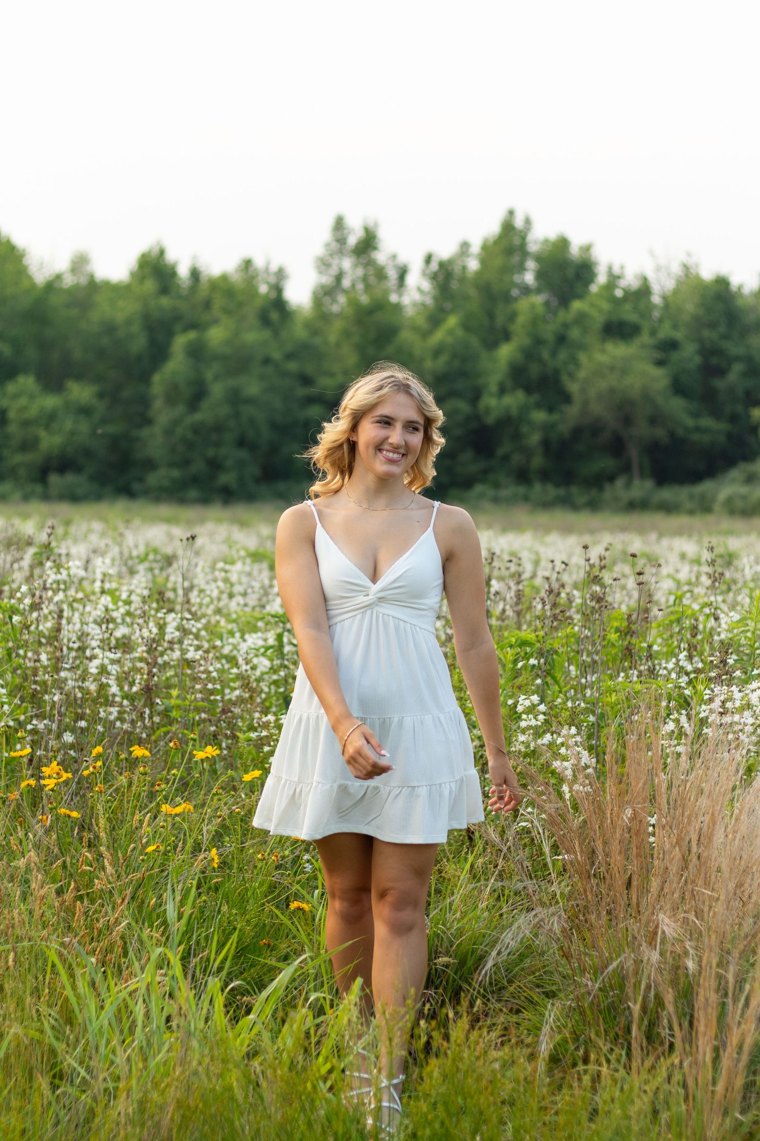 Graduate in white sundress standing in summer wildflower meadow.