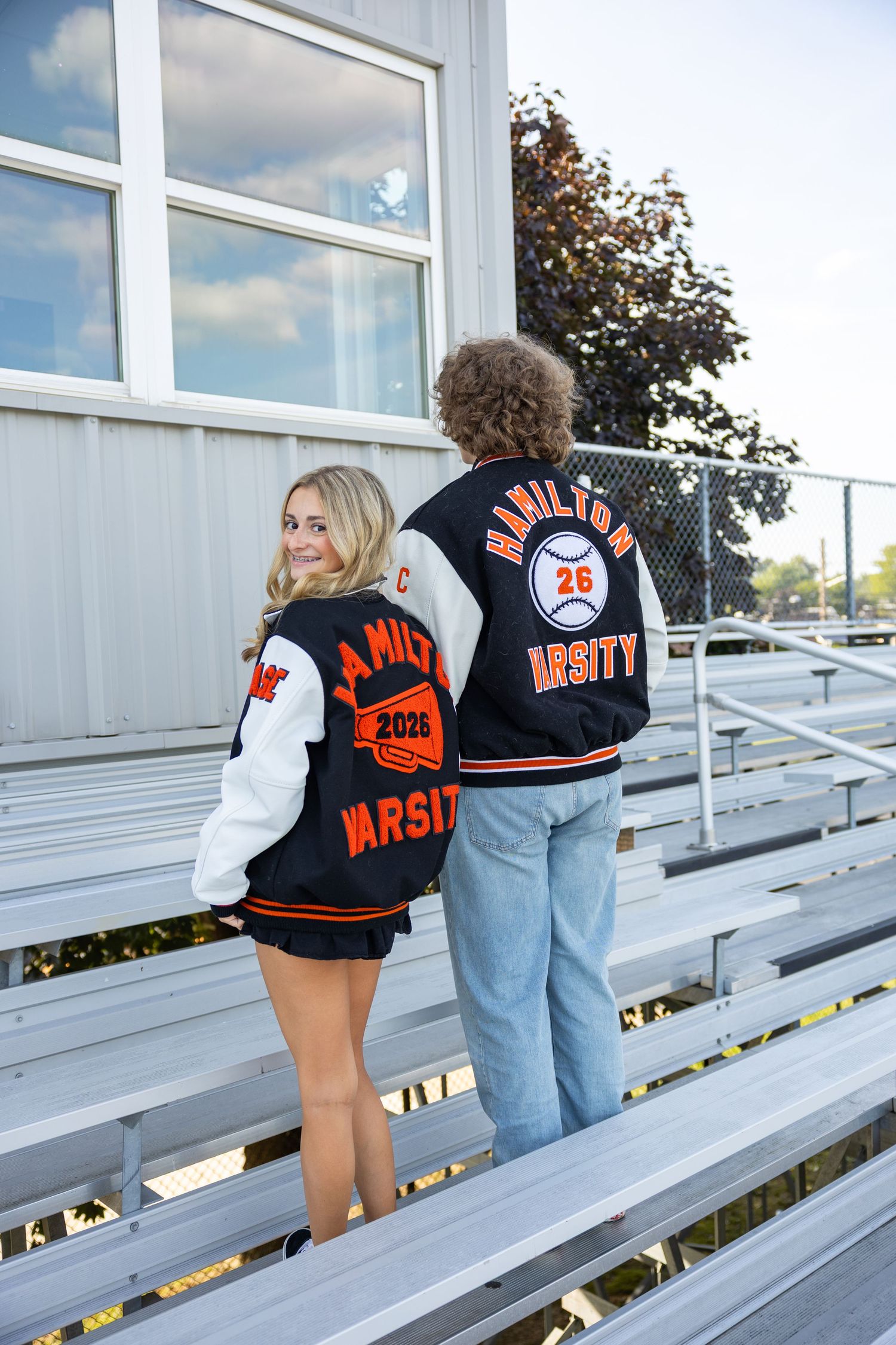 Students in varsity jackets sitting on bleachers outside school building.