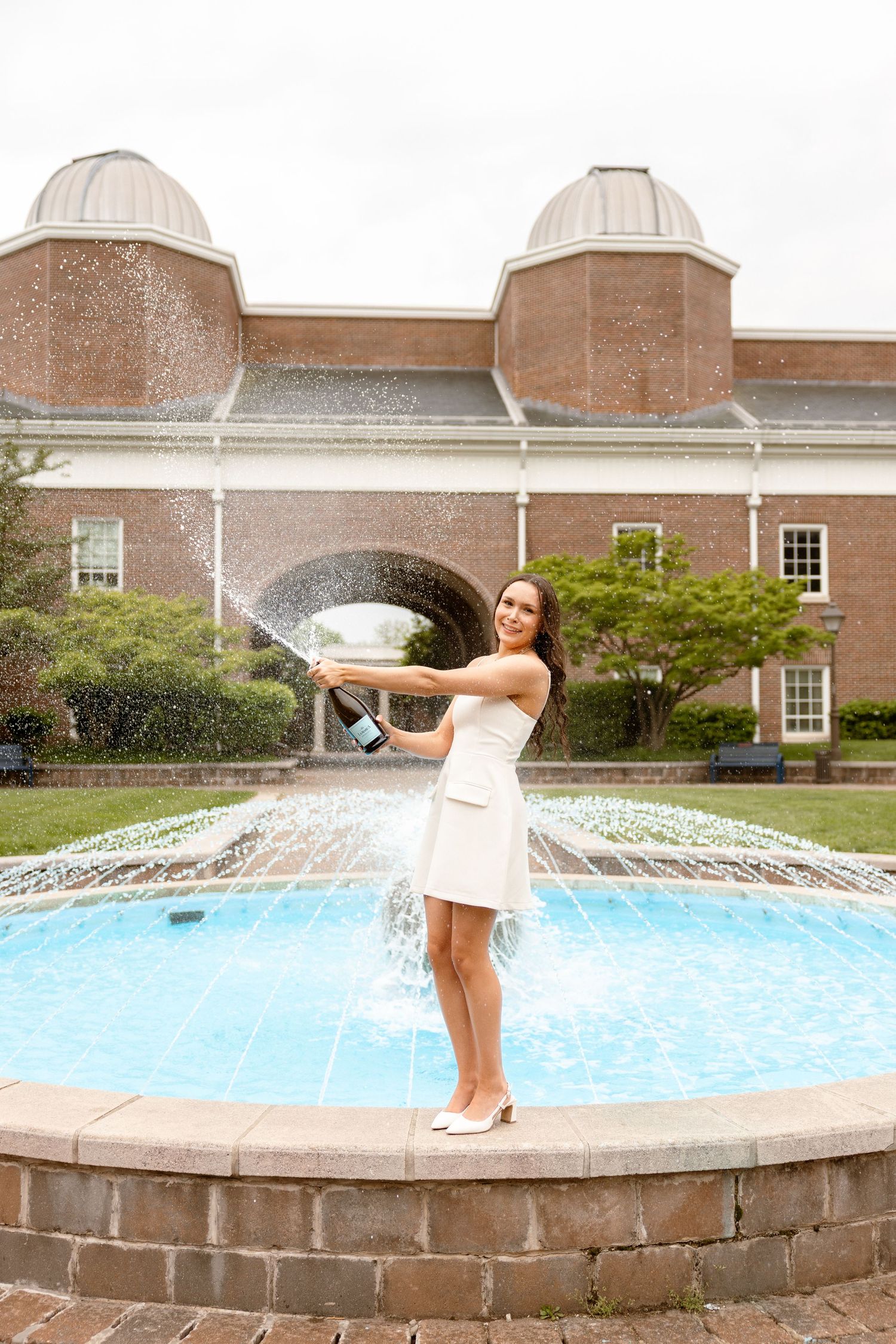 Woman in white dress spraying champagne while standing on a fountain ledge outside a brick building.