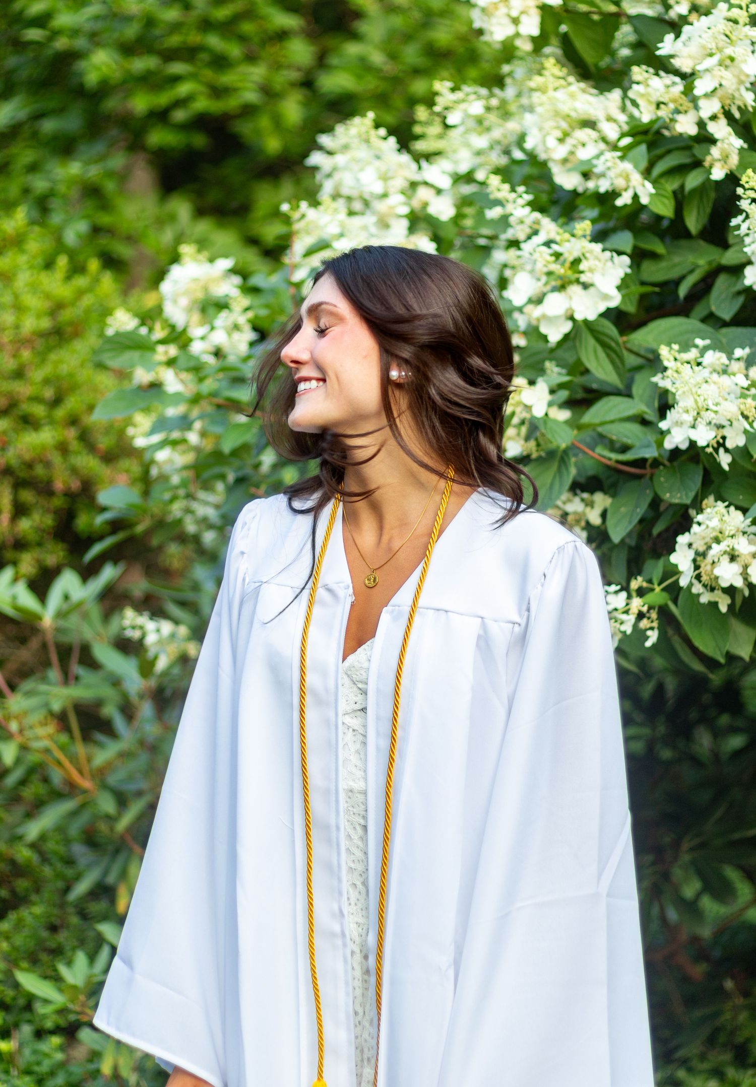 Graduate in white cap and gown smiles near blooming white flowers.