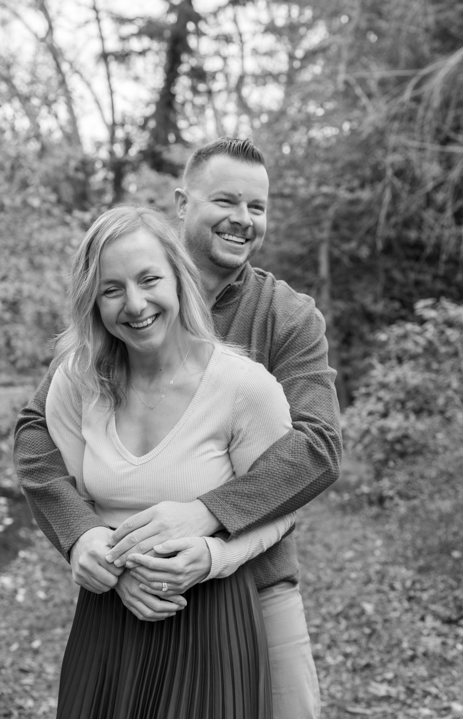 Black and white photo of a smiling couple embracing outdoors in a wooded setting, woman in front with man hugging her from behind.