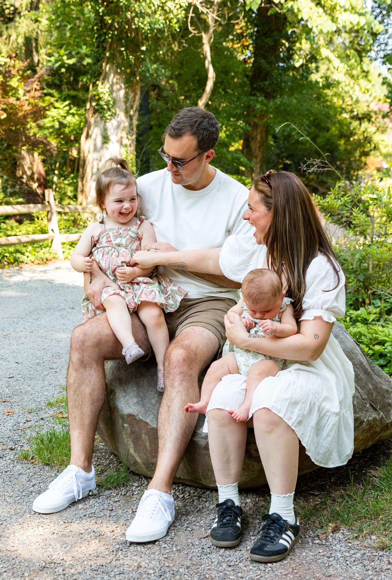 Happy family of four sitting on a rock in a lush green park, parents smiling with toddler girl and newborn baby.