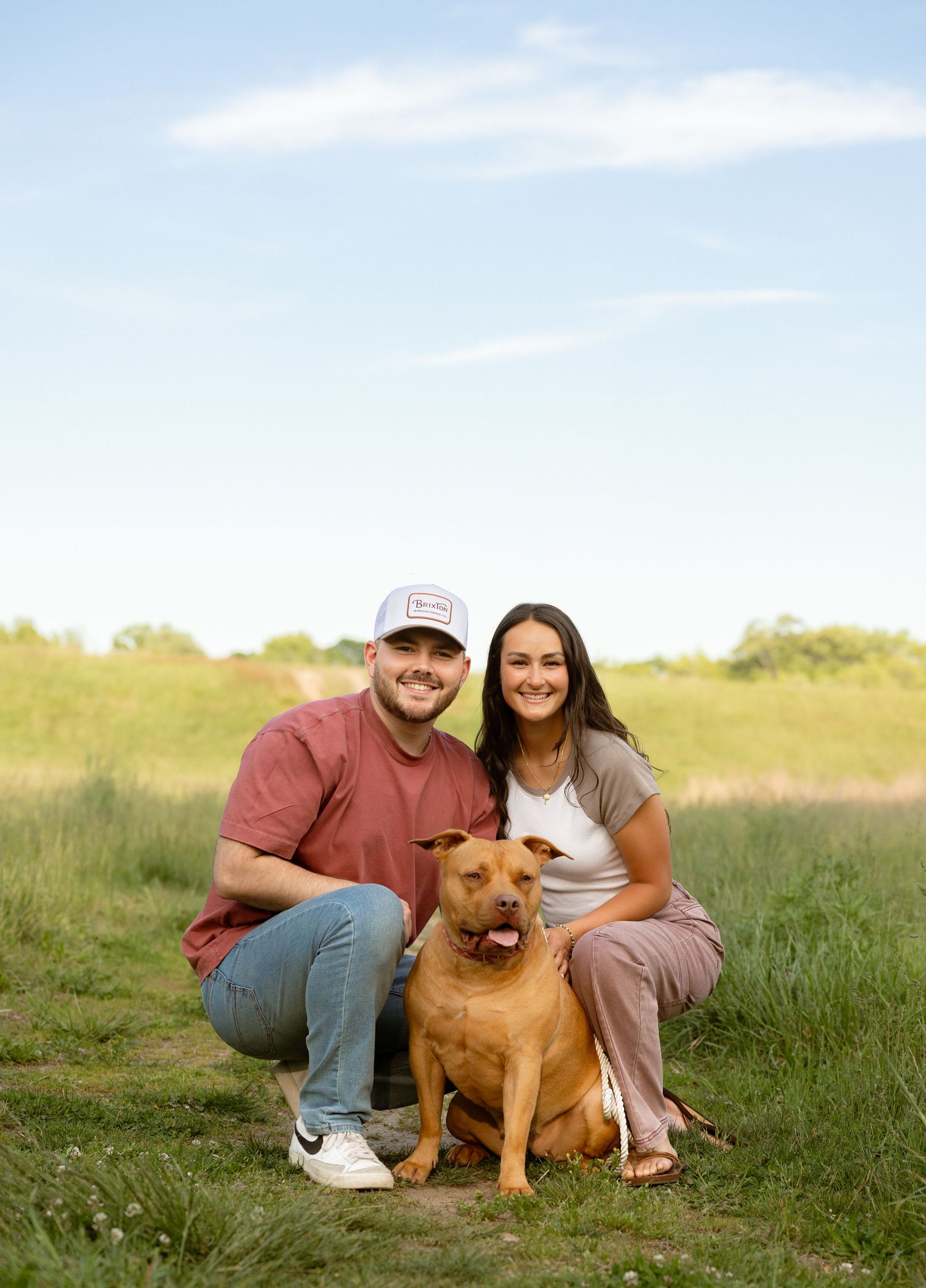 A smiling couple crouches in a green field with their brown dog on a sunny day.