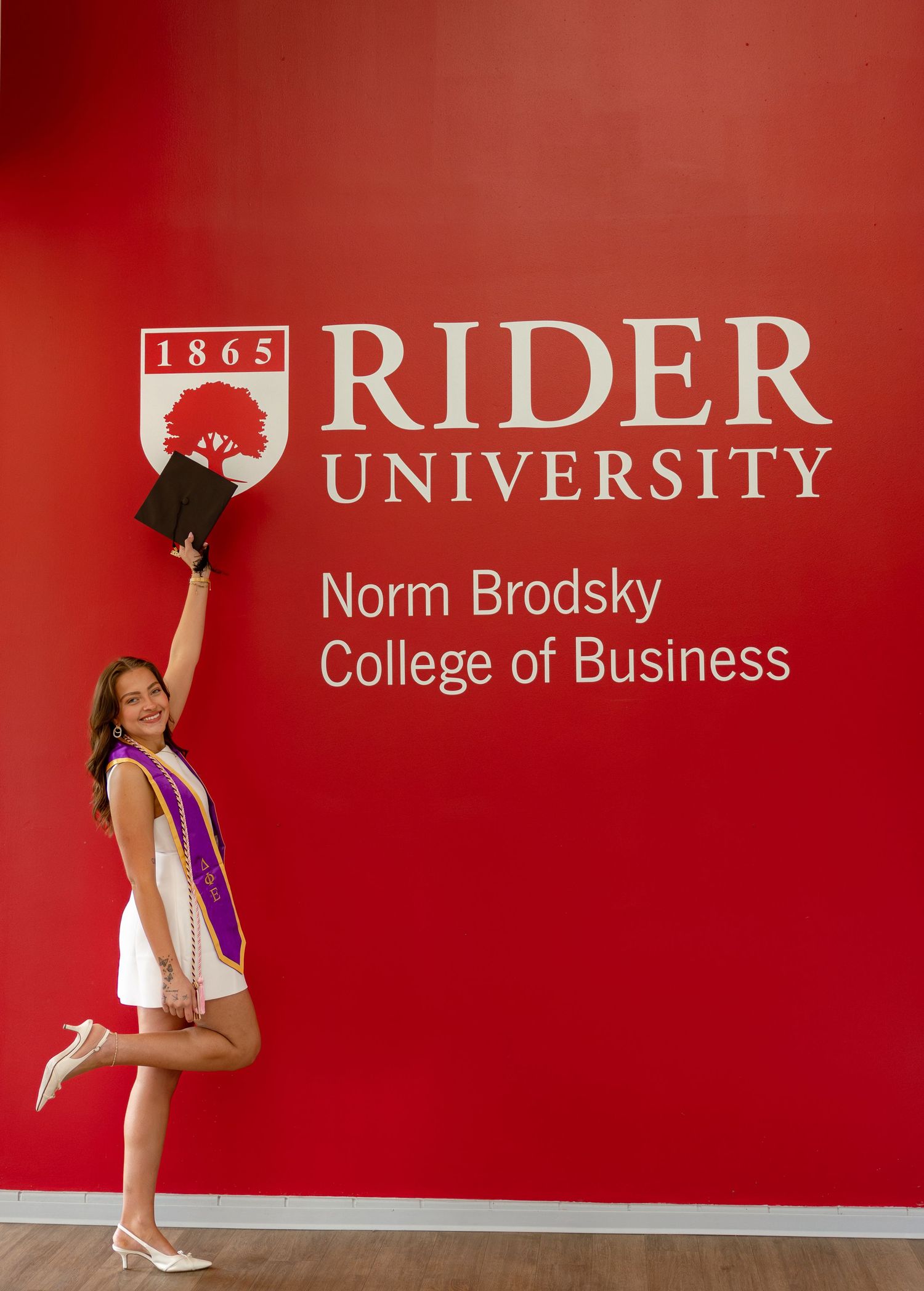 Graduate celebrates at Rider University Norm Brodsky College of Business, holding diploma aloft in white dress with purple sash.