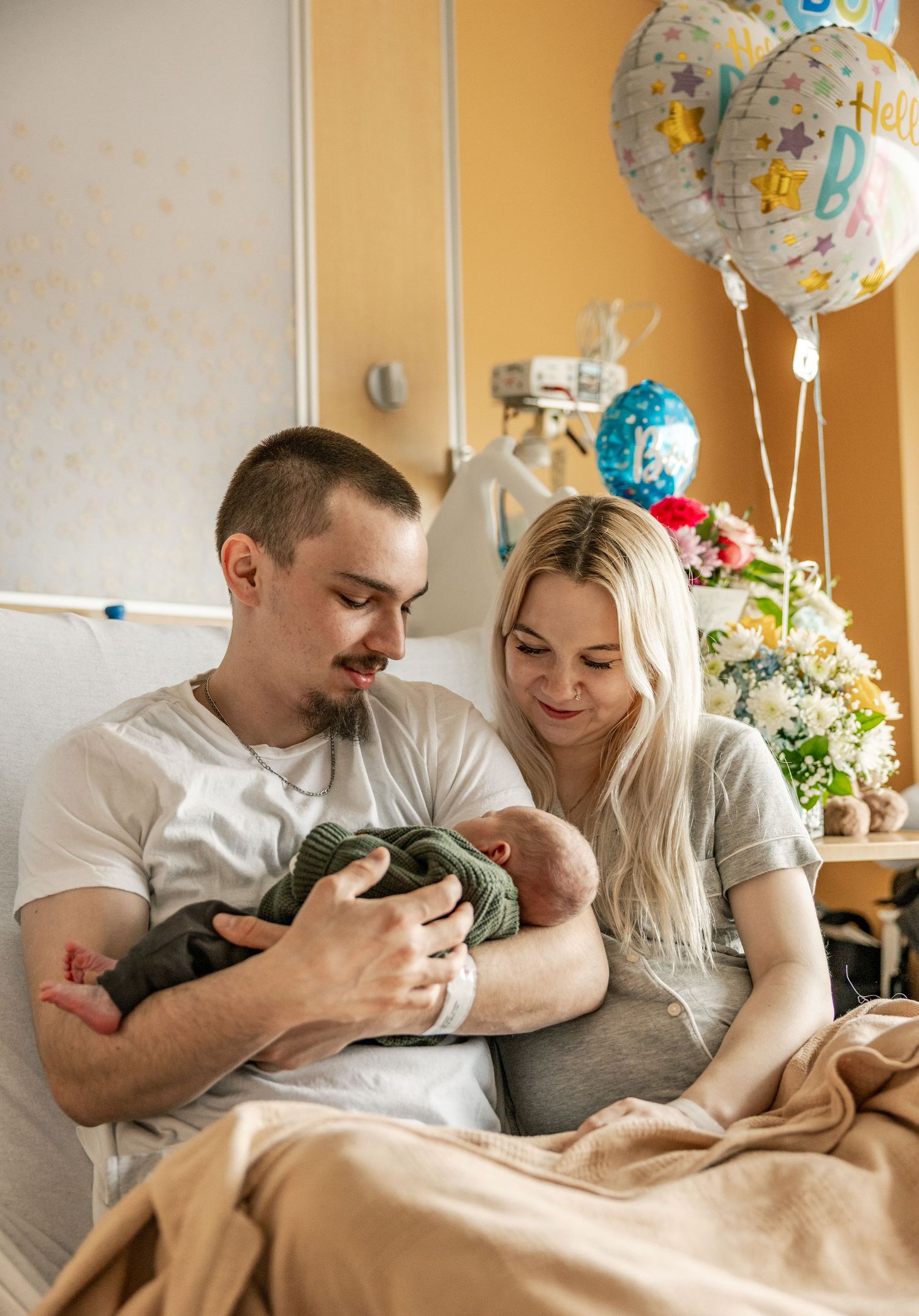 New parents admire their newborn baby in a hospital room decorated with celebration balloons and flowers.