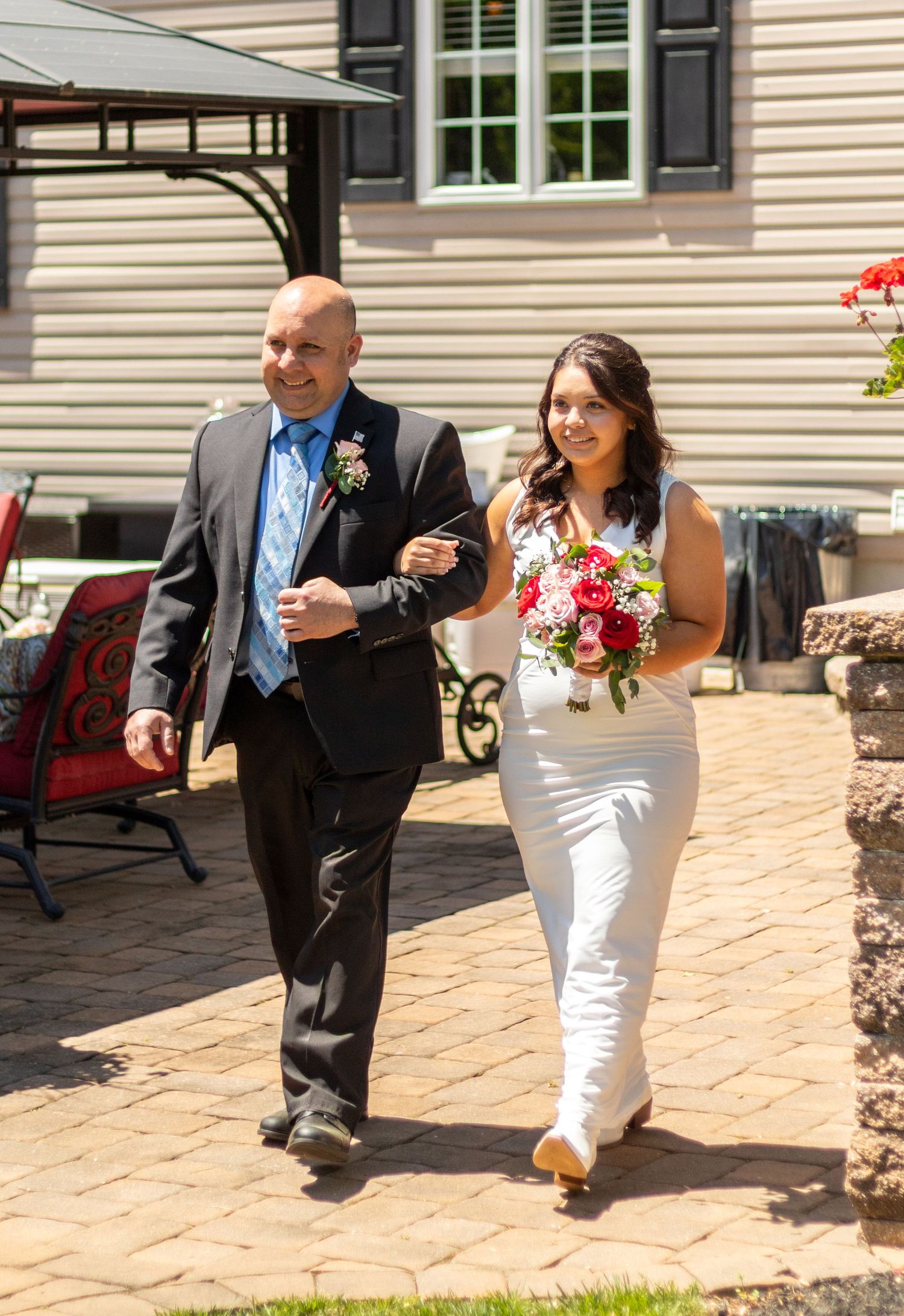 Wedding party members walk along a brick patio outside a beige house.