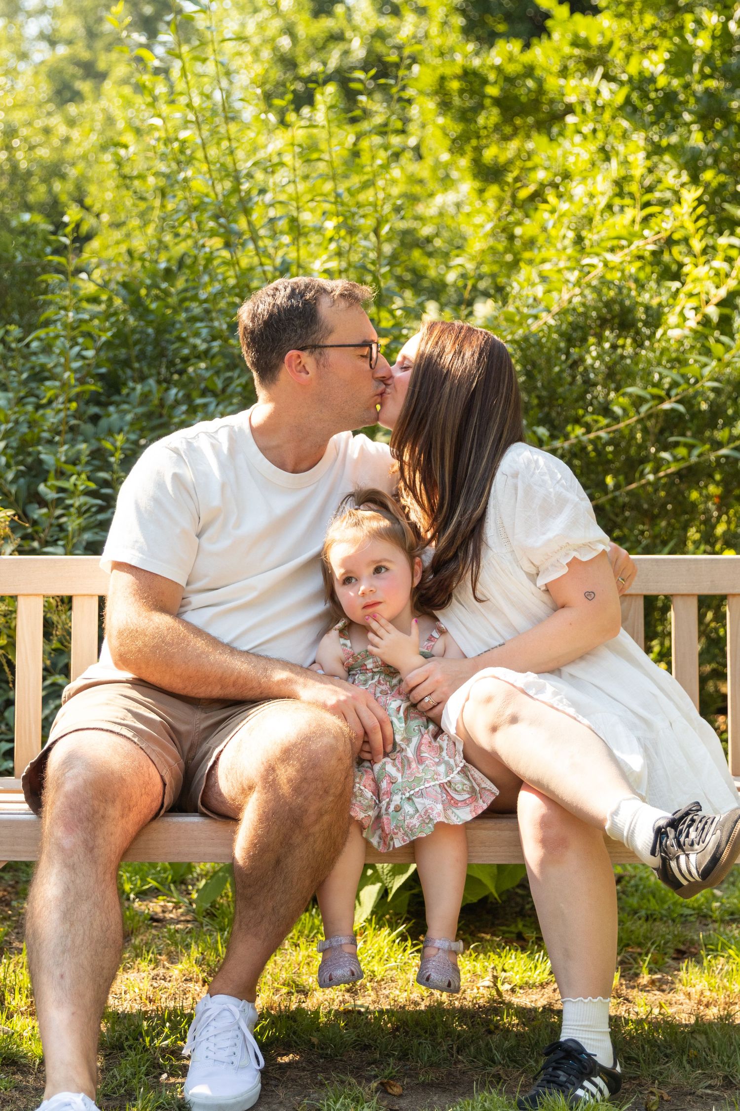 A family shares intimate moments while sitting together on a wooden bench in a sunlit garden.